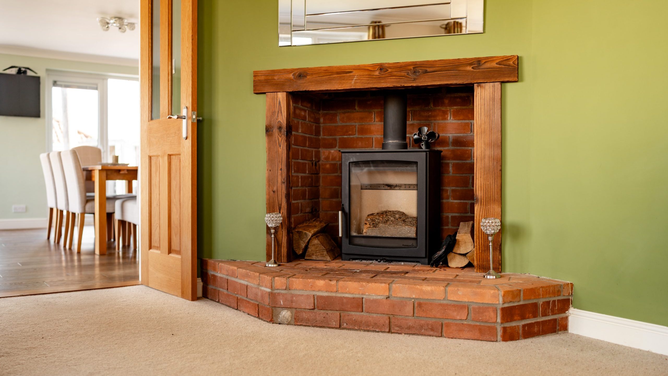 A cozy living room features a wood-burning stove set within a brick fireplace against a green wall. A wooden beam tops the fireplace. To the left, a wooden door opens to a dining area with a table and white chairs. Tan carpet covers the floor.
