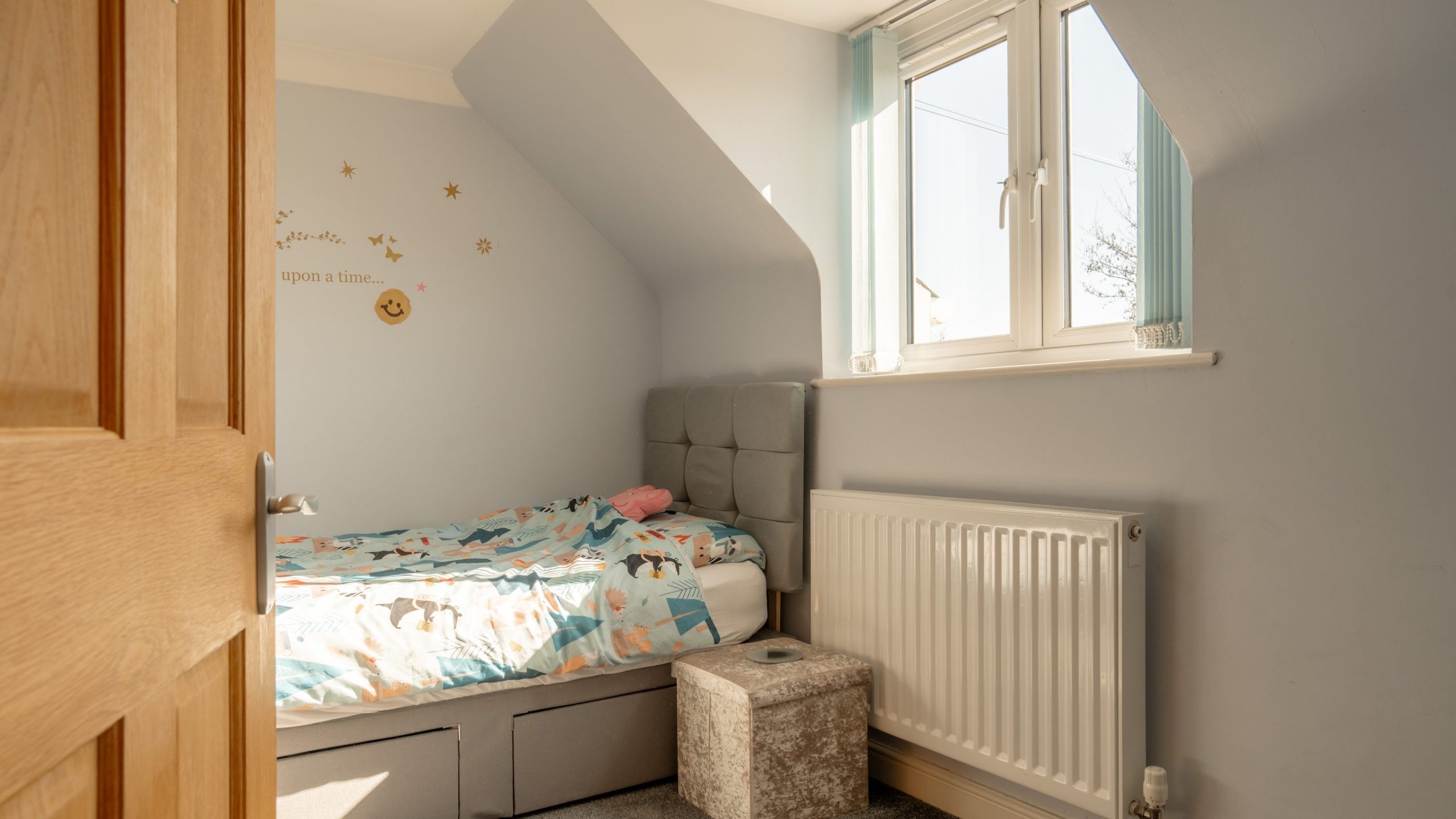 A small, cozy bedroom with a single bed adorned with a colorful children's duvet. The room has light blue walls, a radiator beneath a window with natural light, and a small cushioned ottoman nearby. Wall decor features stars and the phrase