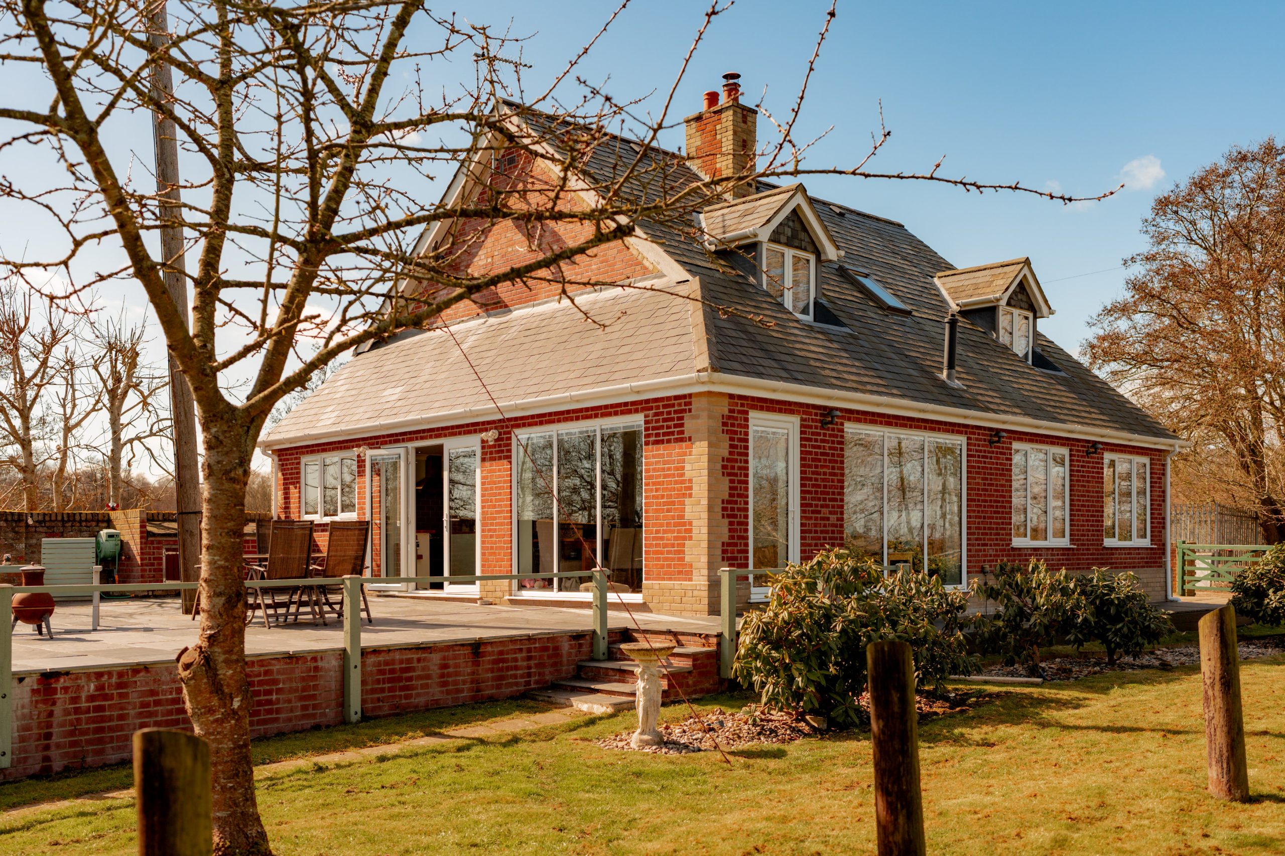 A brick house with a multi-gabled roof, large glass doors, and windows. The house features a wooden deck with outdoor furniture. In the foreground, there's a lawn and a leafless tree under a clear blue sky.