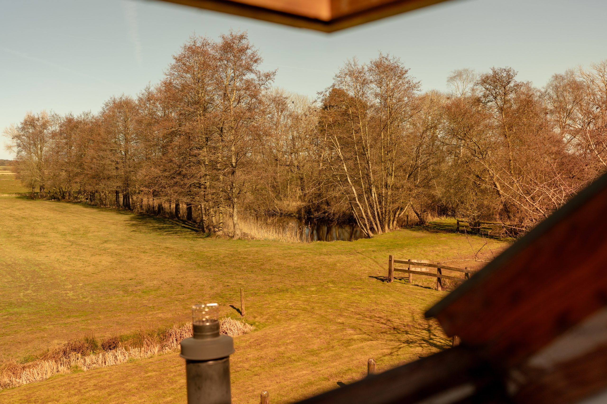 A scenic view of a rural landscape from an attic window. A grassy field stretches toward a row of trees near a pond, with clear skies above. A fence and chimney cap are visible in the foreground.