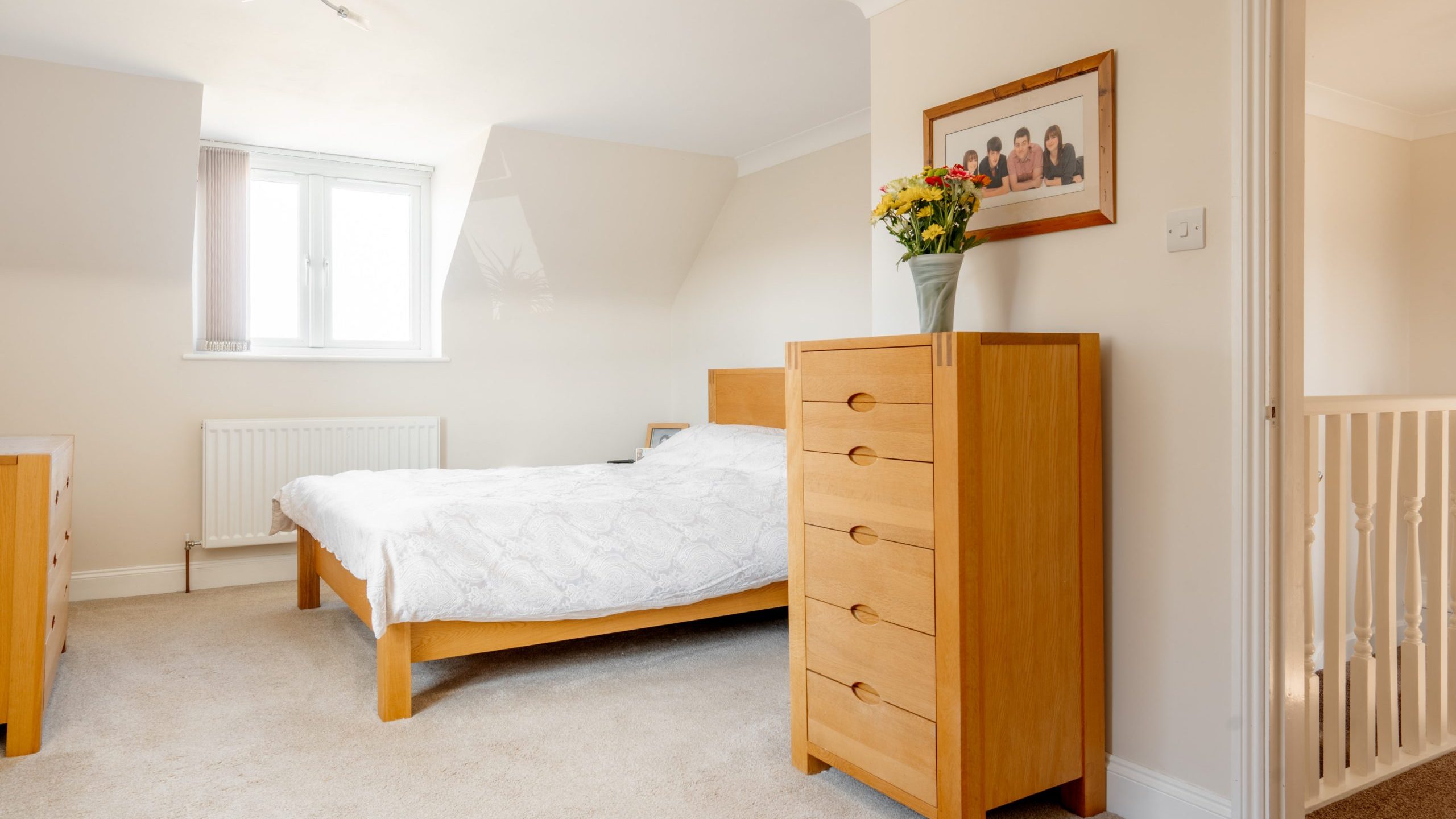 Bright bedroom with a double bed, wooden dresser, and matching bedside table. A vase with flowers is on the dresser. A framed photo hangs on the wall. The room has beige carpet and light walls, and a window letting in natural light.