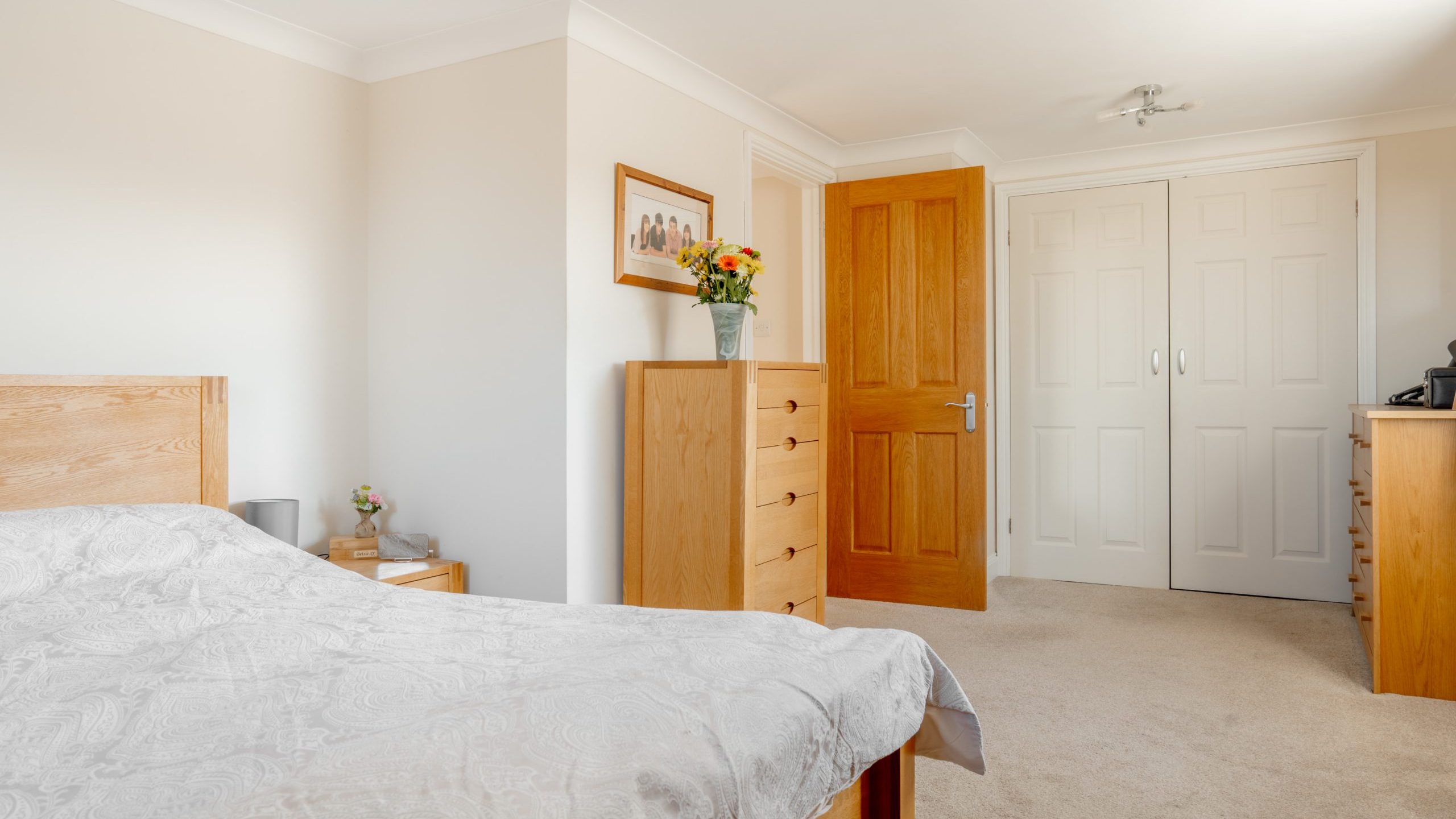 A bright bedroom with light-colored walls features a wooden bed, dresser, and bedside table. A framed picture hangs on the wall, and a vase with flowers sits on the dresser. The room has a closed wooden door and a white closet door.