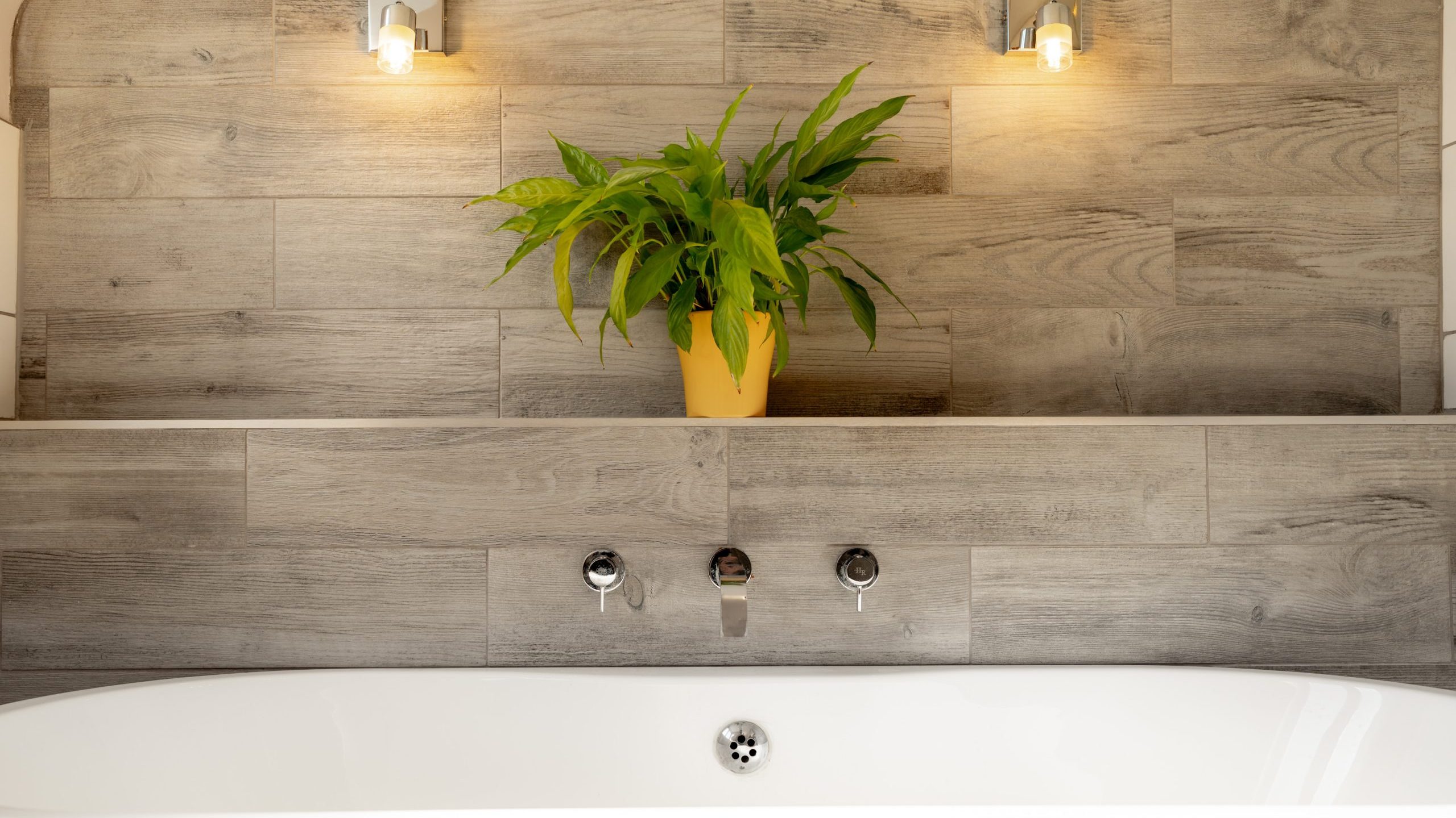 Modern bathroom with a white tub against a textured wooden wall. Above the tub, a green potted plant sits on a shelf. Two small wall-mounted lights illuminate the area, creating a warm and inviting atmosphere.