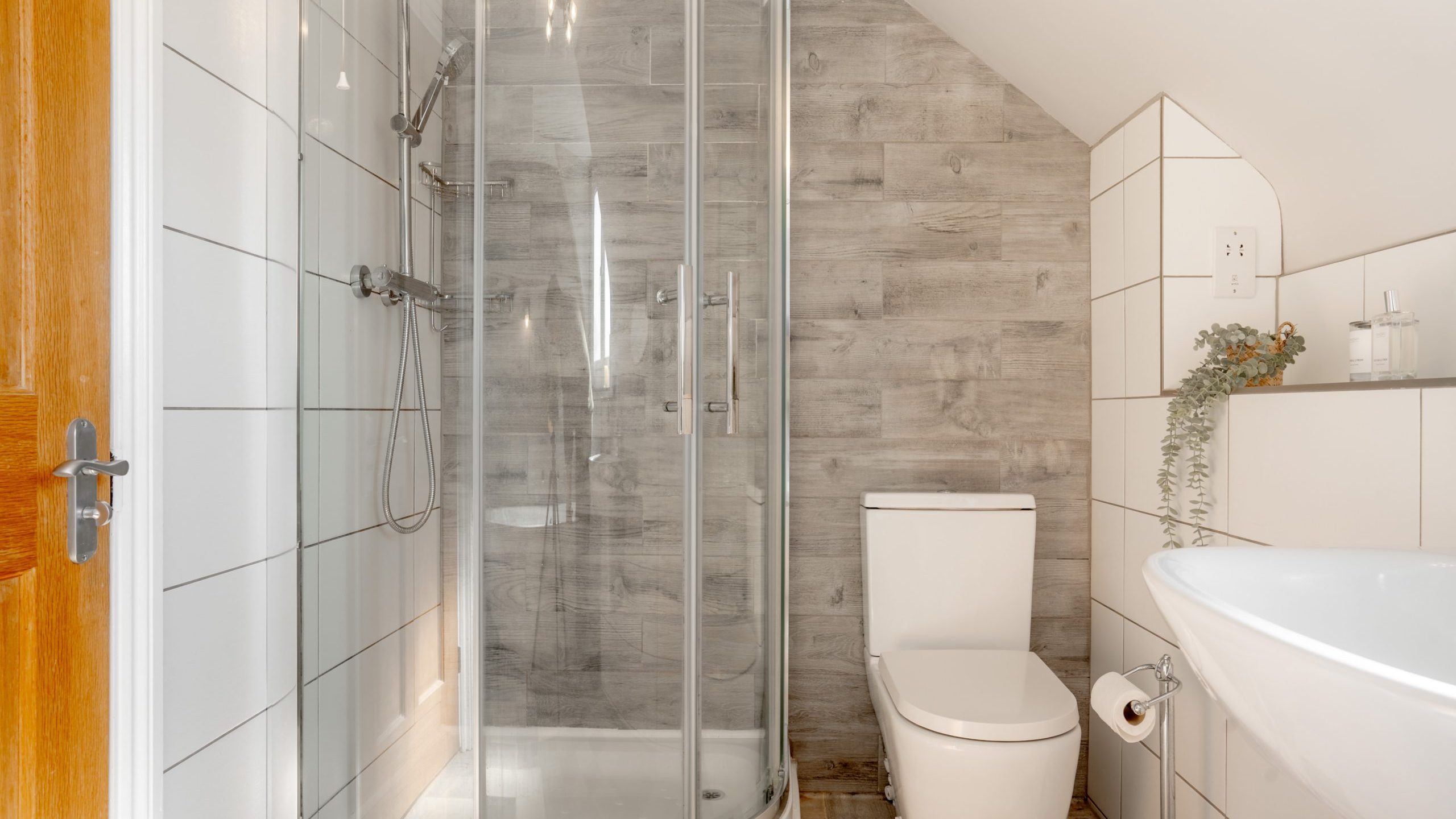 A modern bathroom with a glass-enclosed shower on the left, a white toilet in the center, and a white sink on the right. The walls are tiled in white and wood-like textures. A small plant and toiletries decorate the space.
