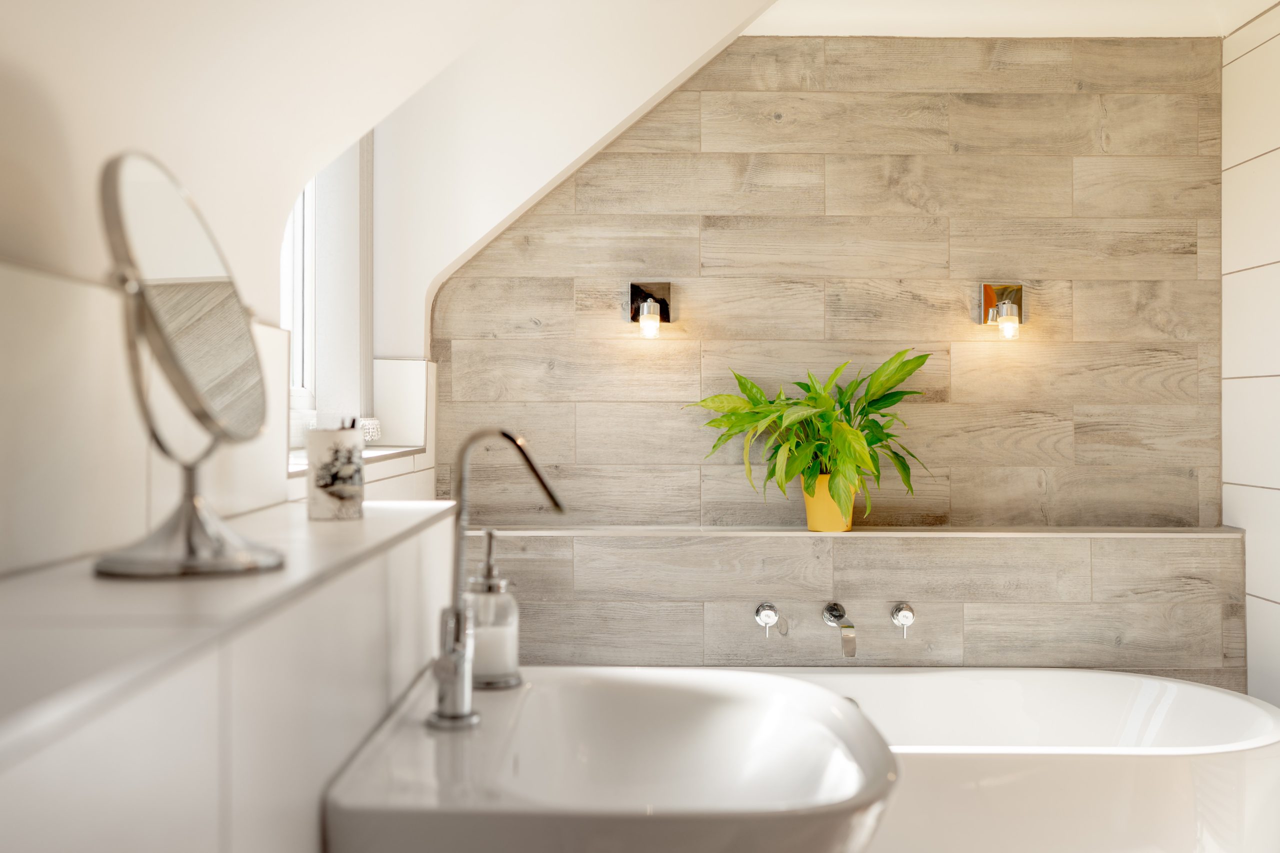 Modern bathroom with a white sink and bathtub. The walls feature light wood-paneling, and two small lights are mounted on the wall. A green plant in a yellow pot sits on a shelf, adding a touch of color and freshness to the space.
