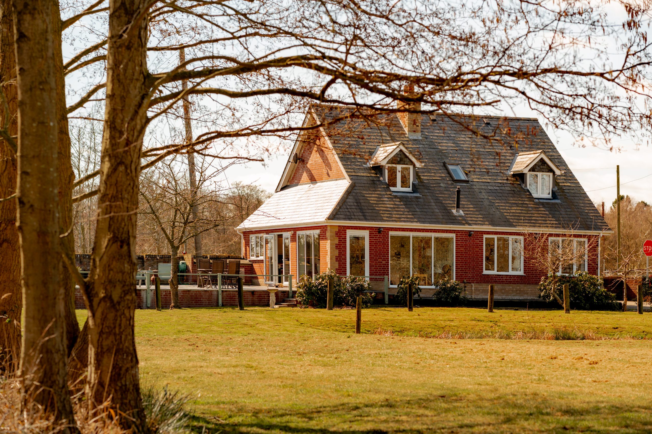 A quaint red-brick house with a steep, shingled roof is nestled among bare trees on a sunny day. The foreground features a grassy lawn and a couple of large tree trunks, and there are people on the patio outside the house.