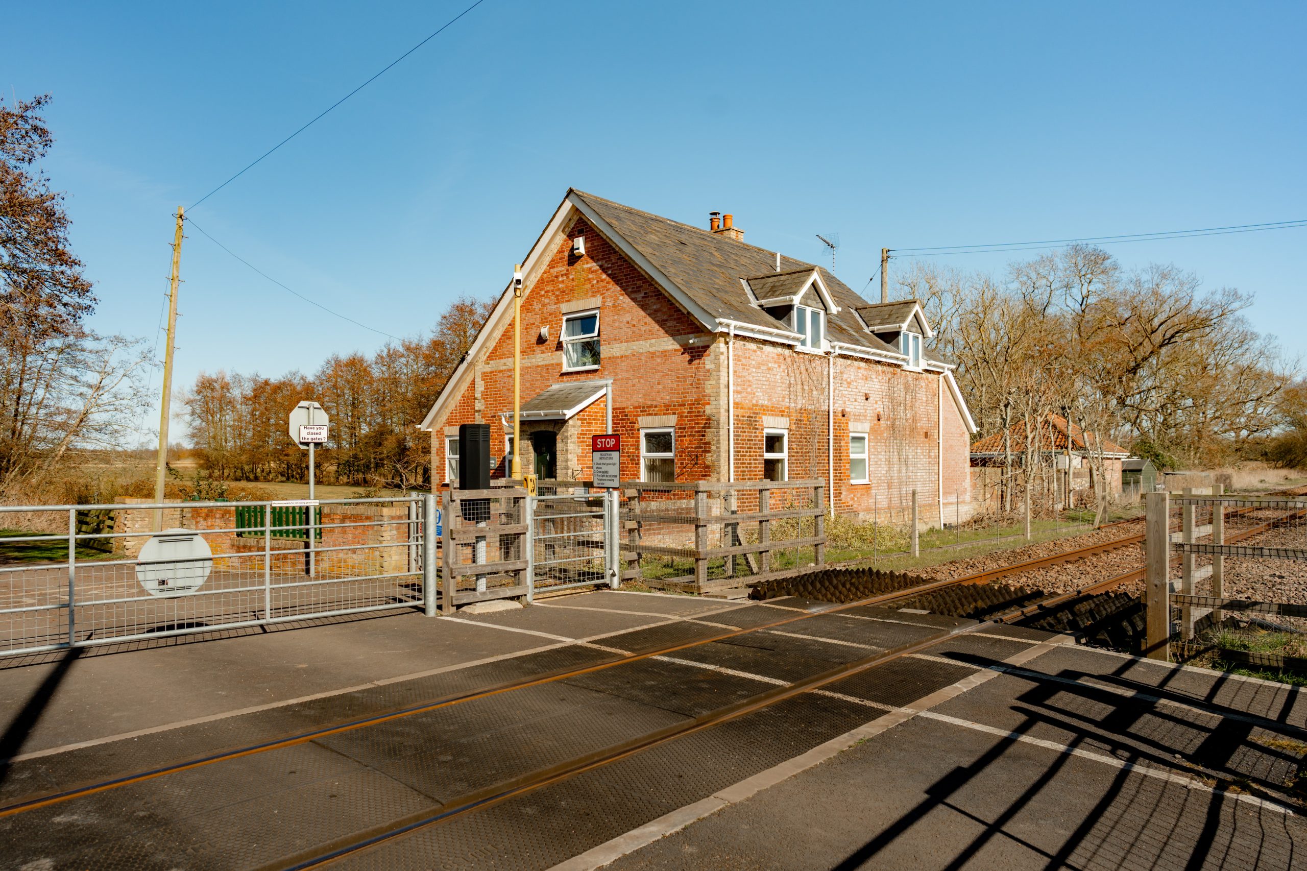 A red brick house stands beside a railroad crossing. The area is surrounded by leafless trees and a clear blue sky. The crossing features barriers, tracks, and a warning sign. The scene is lit by bright sunlight.