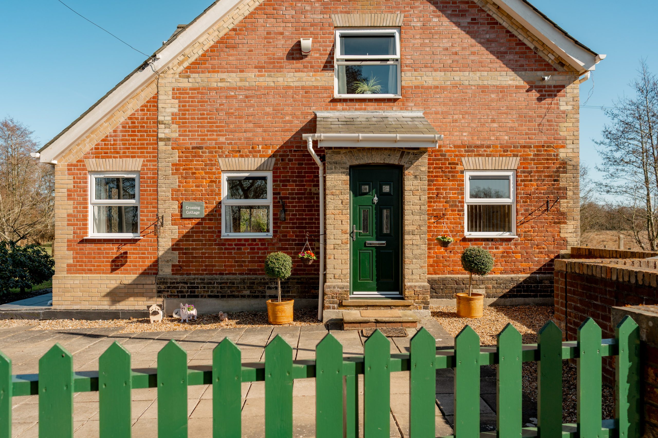 A red brick house with a green wooden fence in front. The house has a green front door, two windows on either side, and is surrounded by a neat garden with potted plants. Blue sky and trees are visible in the background.