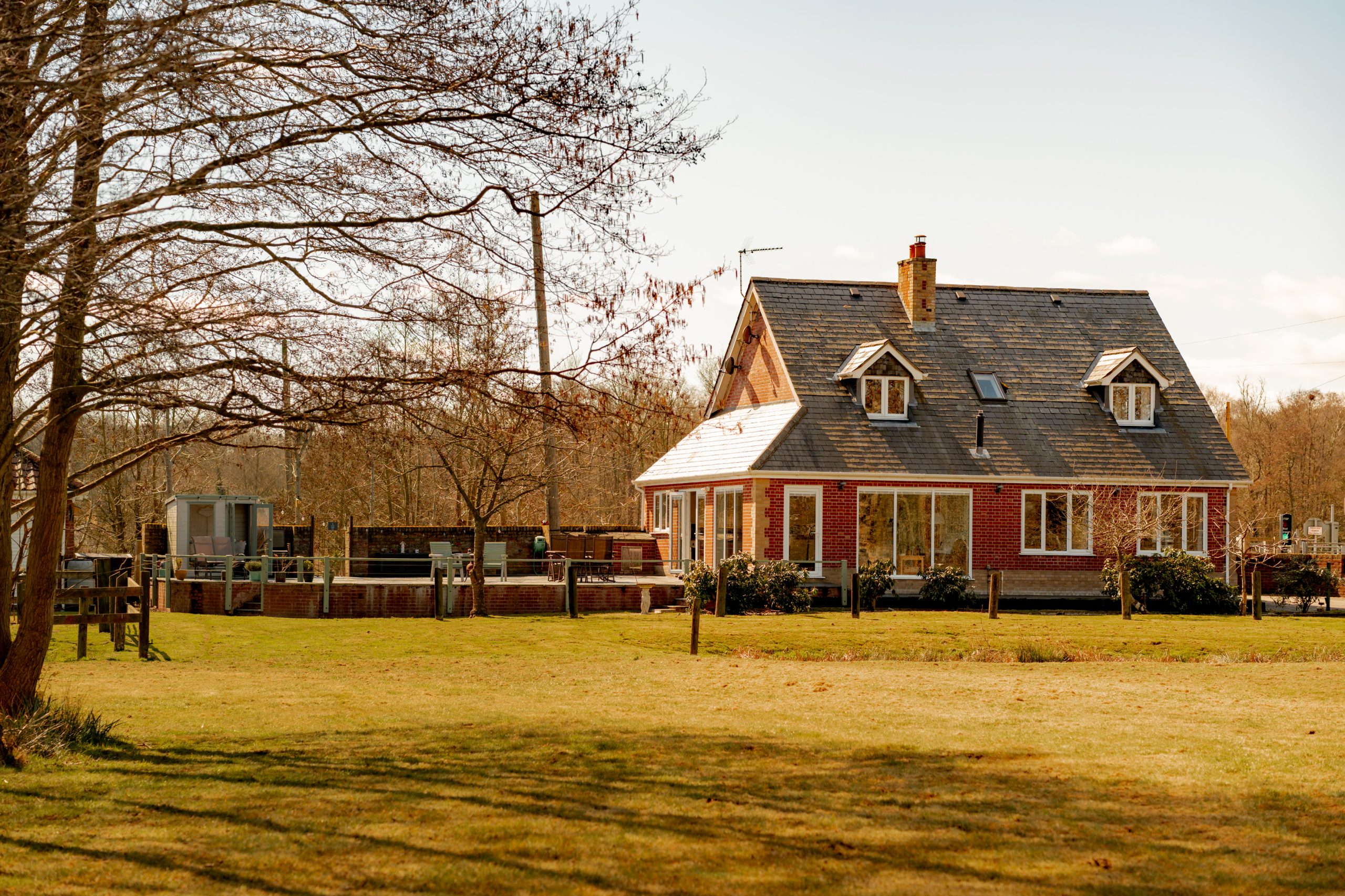 A cozy red-brick house with a steep gray roof and multiple windows sits in a sunlit yard. Leafless trees surround the property on a clear day. A fenced area and outbuilding are visible in the background.