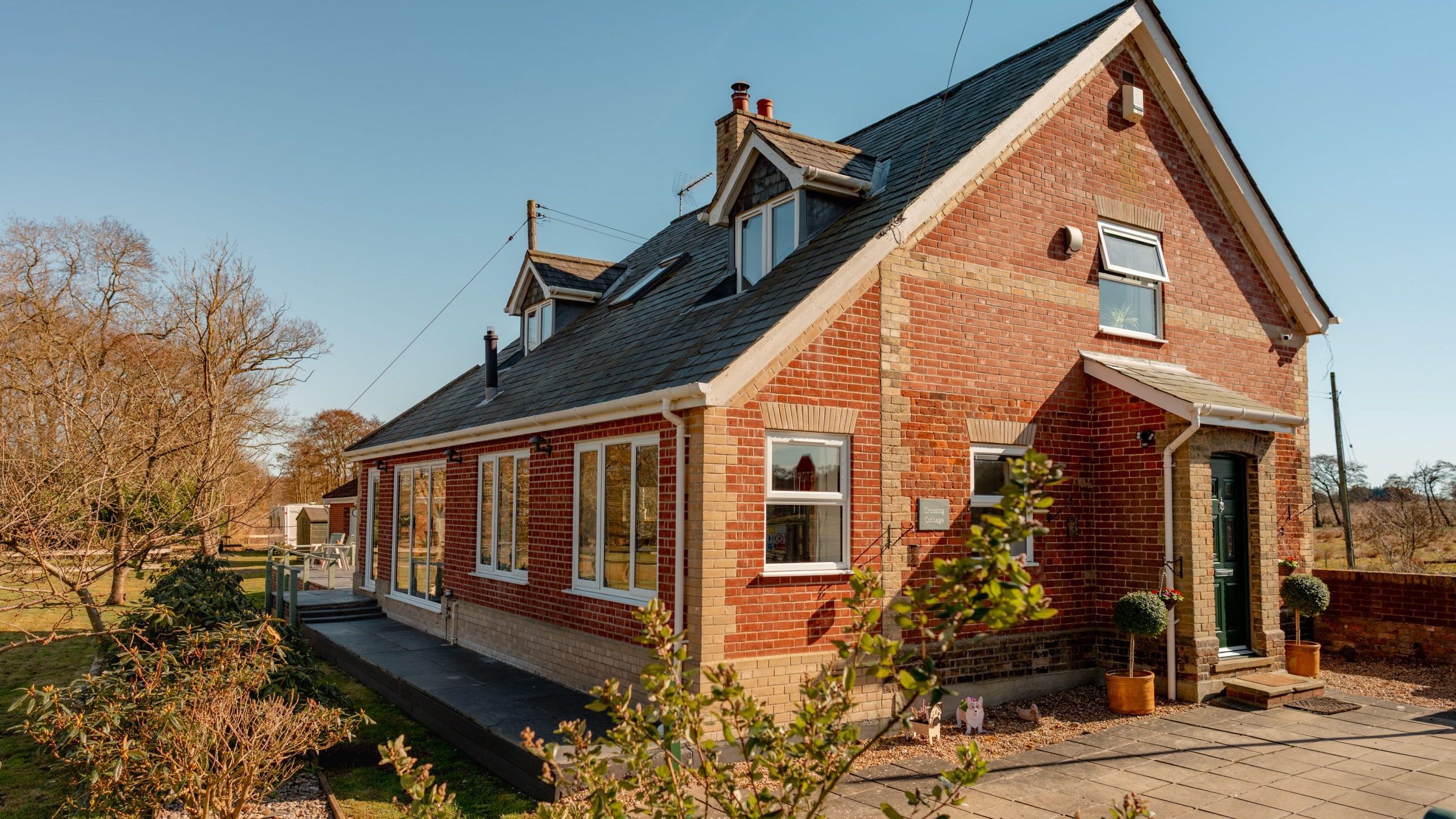 A charming red brick house with a dark gray roof. It features multiple windows and a glass door. The house is surrounded by a well-maintained garden with bushes and trees, set against a clear blue sky.