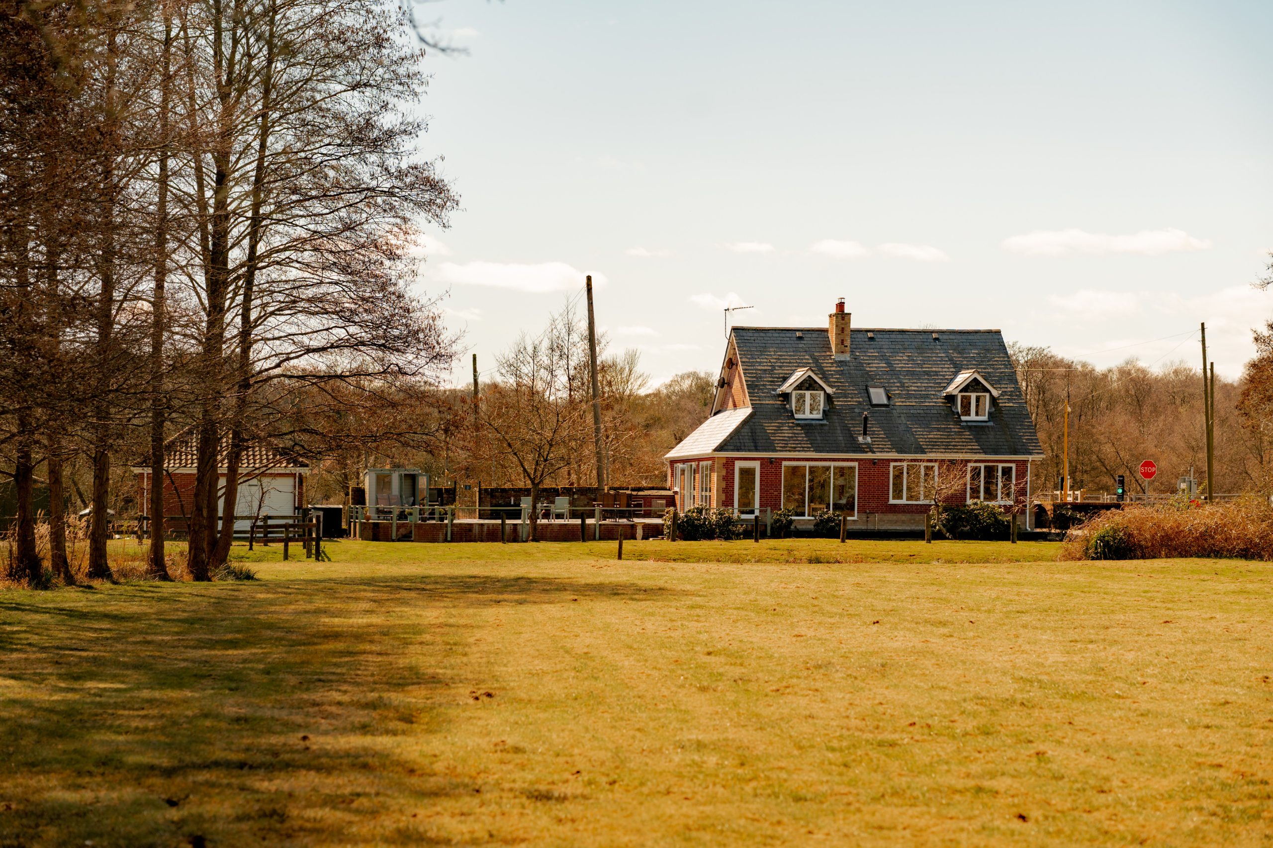 A charming country house with a slate roof stands surrounded by bare trees and a grassy field under a clear sky. A wooden fence encloses the property, and a small outbuilding is nearby. Sparse clouds dot the blue sky.