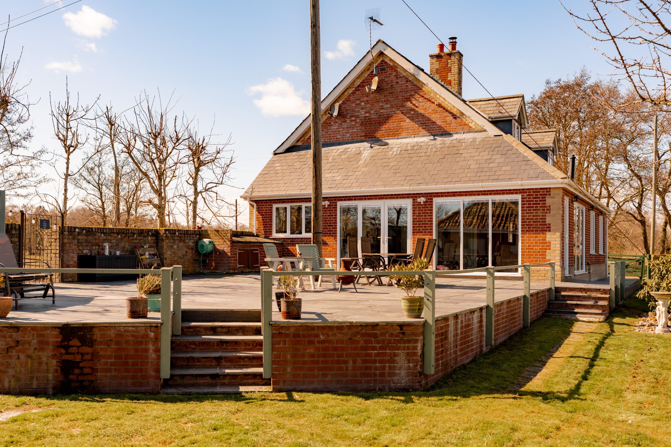 A red brick house with large windows and a gabled roof, surrounded by a spacious wooden deck. There are outdoor chairs and tables on the deck, and a grassy lawn with bare trees in the background, under a clear blue sky.