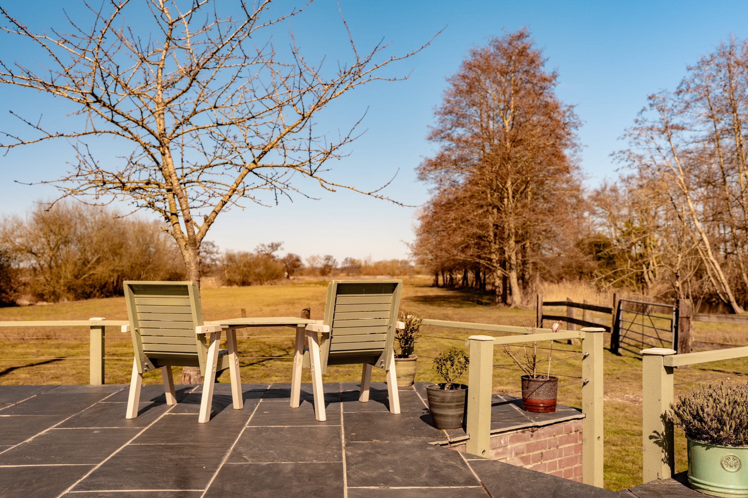 Two wooden chairs and a small table sit on a patio overlooking a peaceful countryside. Leafless trees and a field stretch into the distance under a clear blue sky, creating a serene, rural scene.