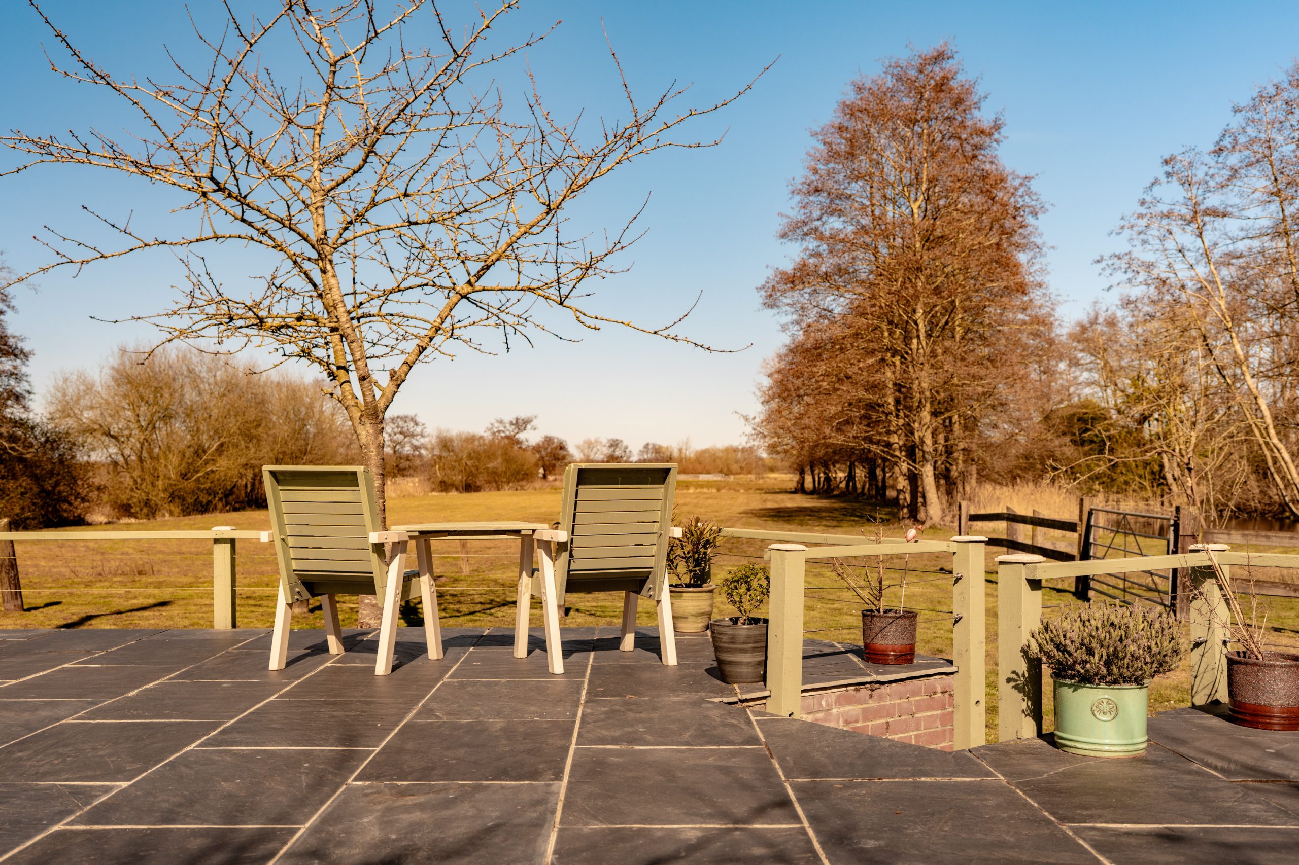 Two wooden chairs on a stone patio overlook a scenic landscape with sparse trees under a clear blue sky. Near the chairs, potted plants add a touch of greenery to the serene outdoor setting.