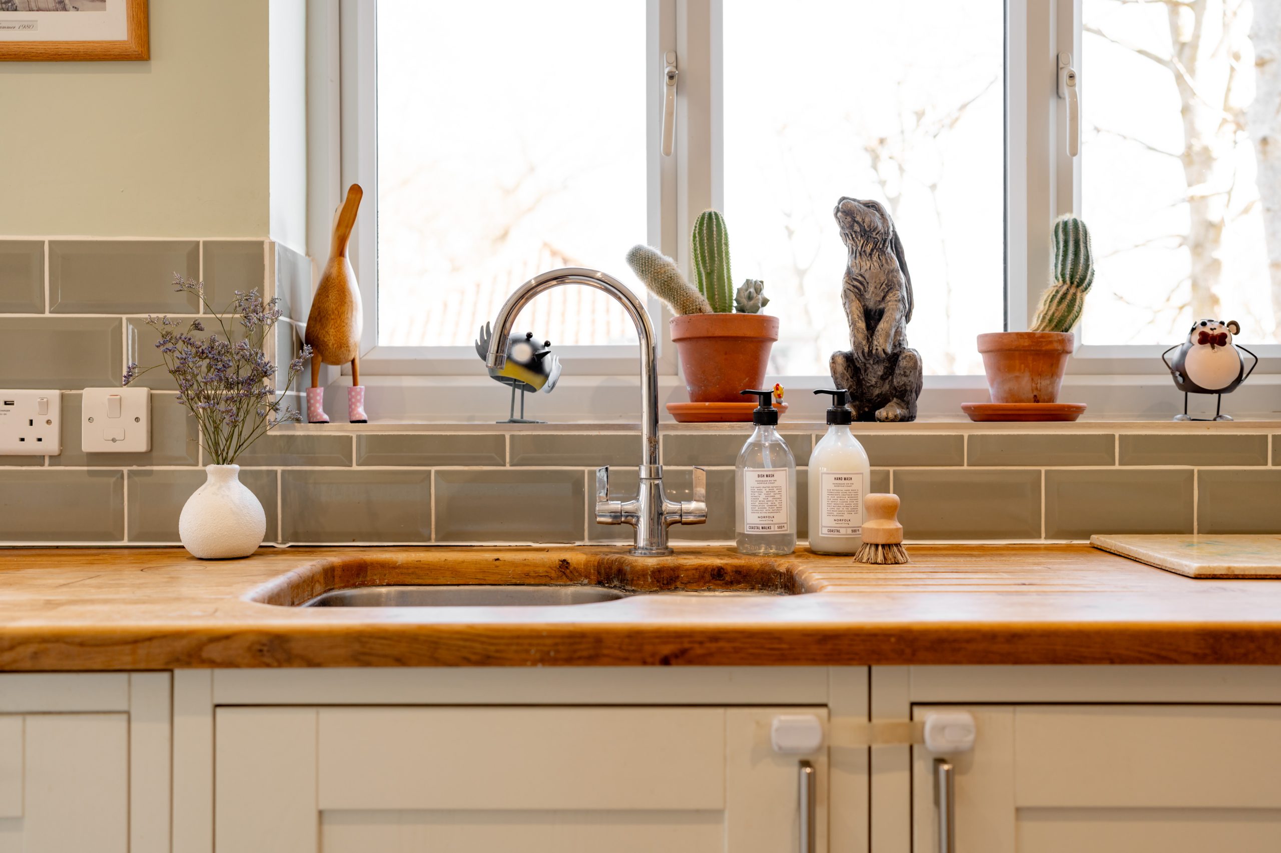 A kitchen sink with a wooden countertop, a silver faucet, and two soap dispensers. Potted cacti and decorative figurines are displayed on the windowsill, with sunlight streaming in from a large window. A small vase with flowers sits on the counter.