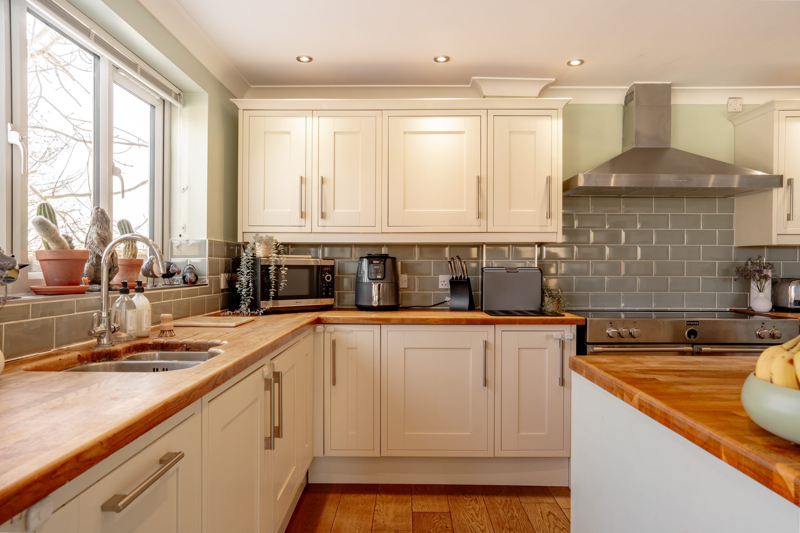 A modern kitchen with wooden countertops, white cabinets, and a tiled backsplash. It features a stainless steel range hood, microwave, toaster, and a coffee maker. A bowl of bananas and potted plants are on the counter near a window.