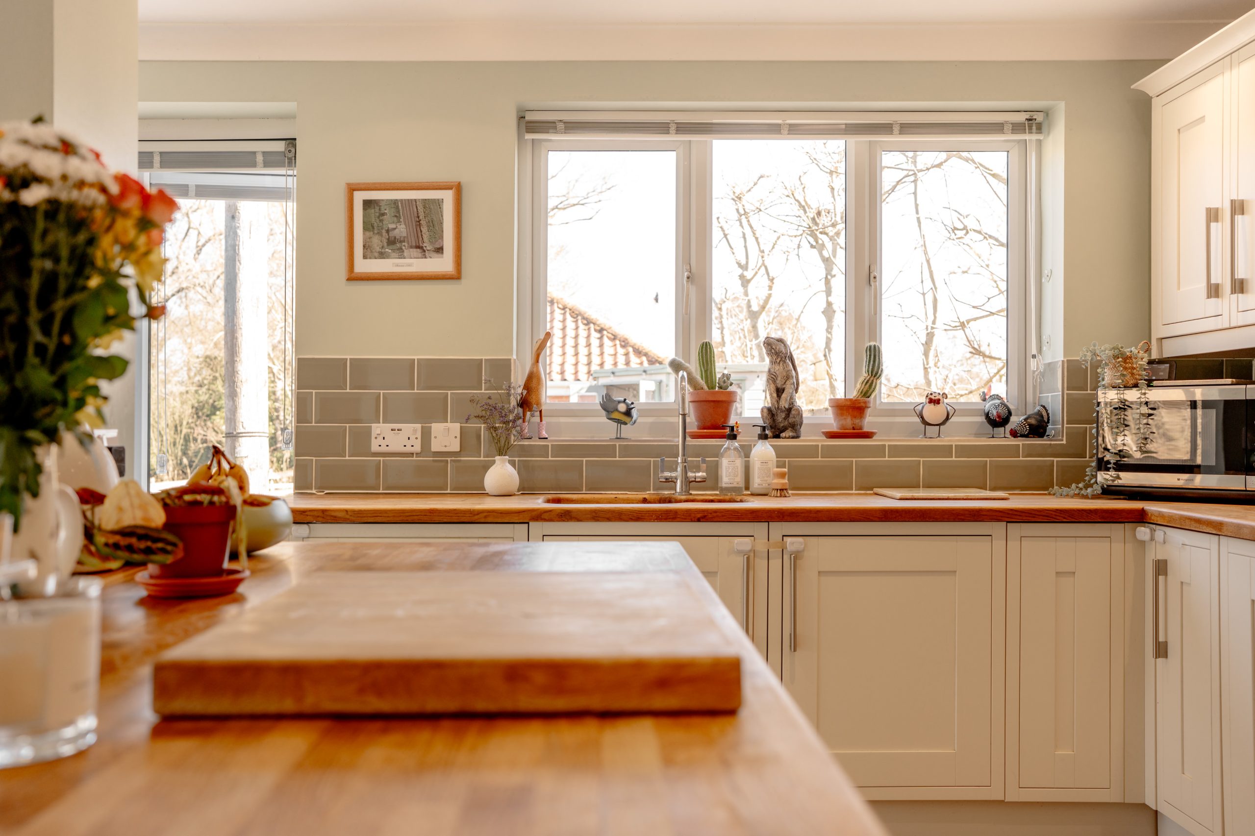 A bright kitchen with white cabinets, a wooden countertop, and a large cutting board in the foreground. Potted plants are placed on the windowsill, and a window offers a view of trees outside. A microwave is visible on the right.