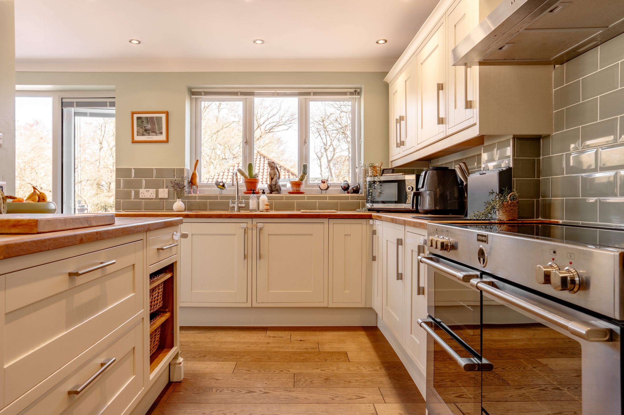 A bright kitchen with wooden floors, white cabinets, and stainless steel appliances. A large window above the sink lets in natural light. A fruit bowl is on the island, and a microwave and kettle sit on the countertop.