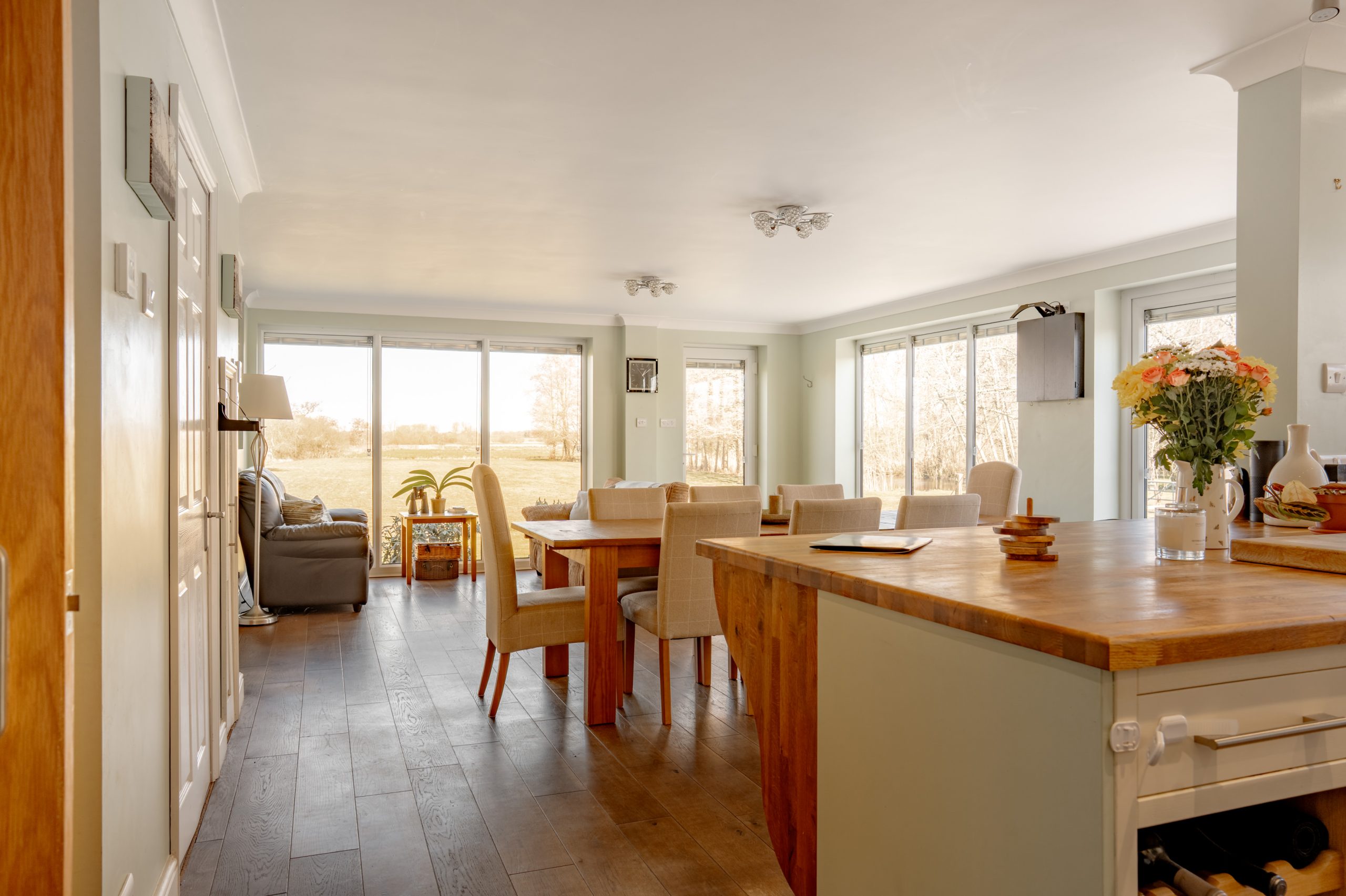 Spacious, sunlit dining area connected to a living room and a kitchen with wooden floors and furniture. Large windows offer a view of the outdoors. A bouquet of flowers and a fruit platter are on the kitchen counter.