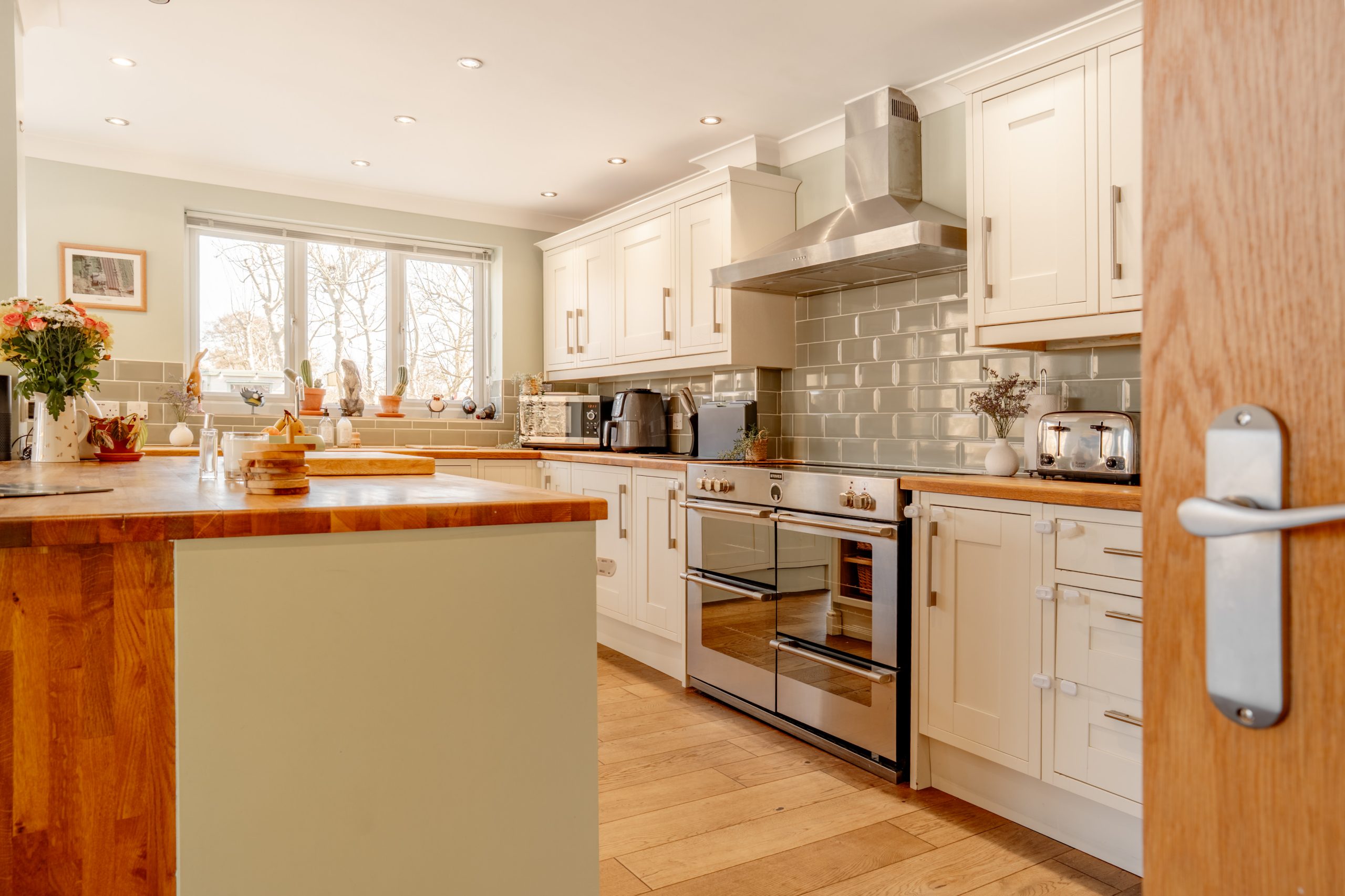 A bright kitchen with white cabinets, a stainless steel range hood, and wooden countertops. There are various kitchen appliances and decor items on the counters. A window lets in natural light, and a bouquet of flowers sits on an island.