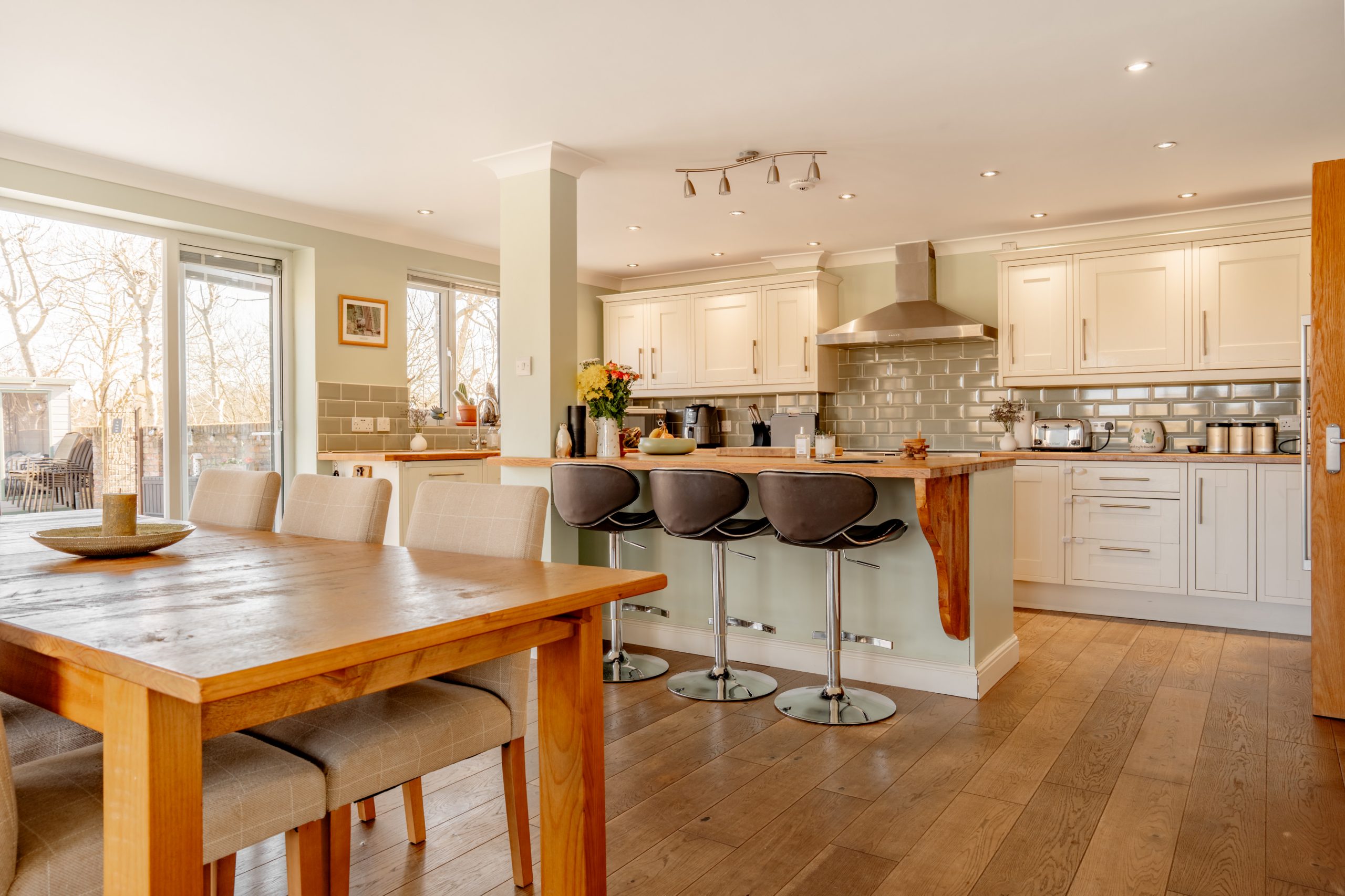 A modern kitchen with white cabinetry and stainless steel appliances, including a range hood. A wooden dining table with beige chairs is in the foreground. Three gray barstools line the kitchen island. Large windows let in natural light.