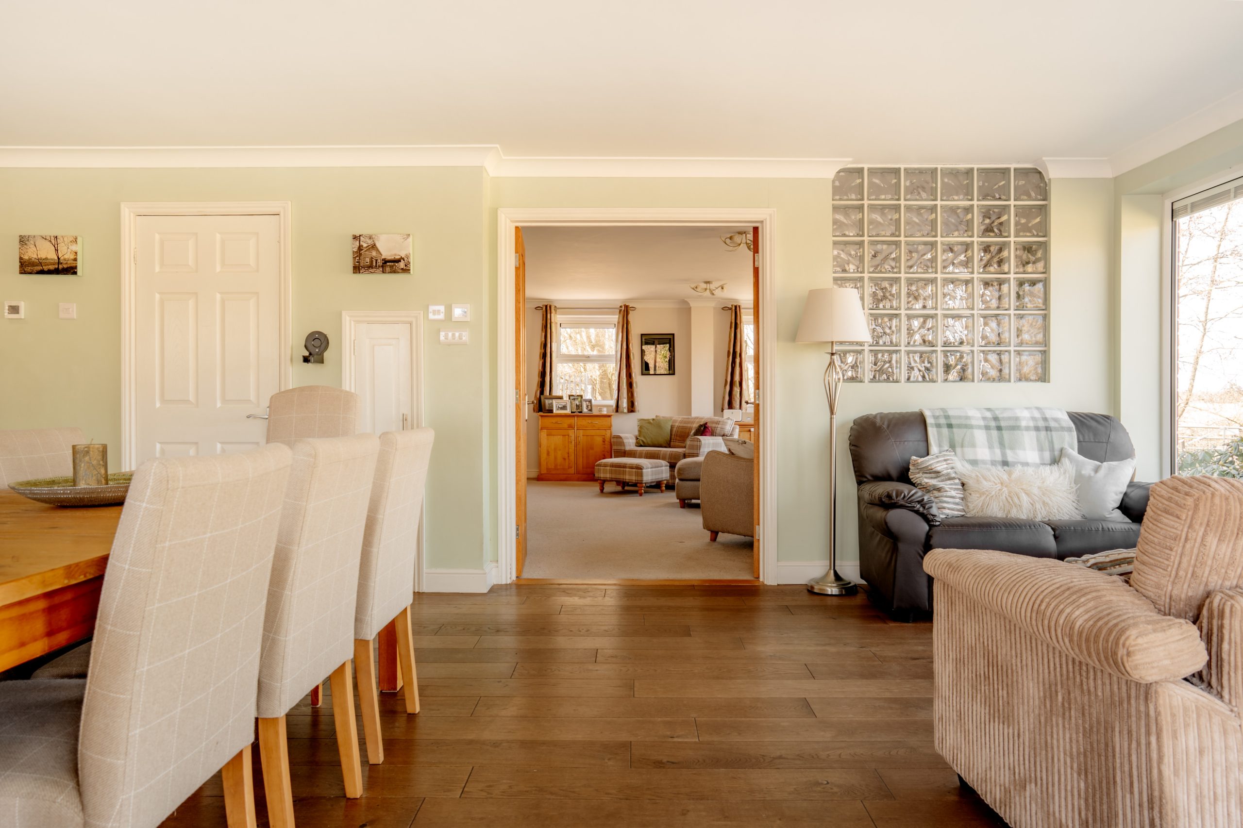 Modern open-plan living area with hardwood floors, featuring a dining table with beige chairs on the left and a cozy sitting area with plush armchairs on the right. A glass block wall accentuates the light-filled space, leading to another living room.
