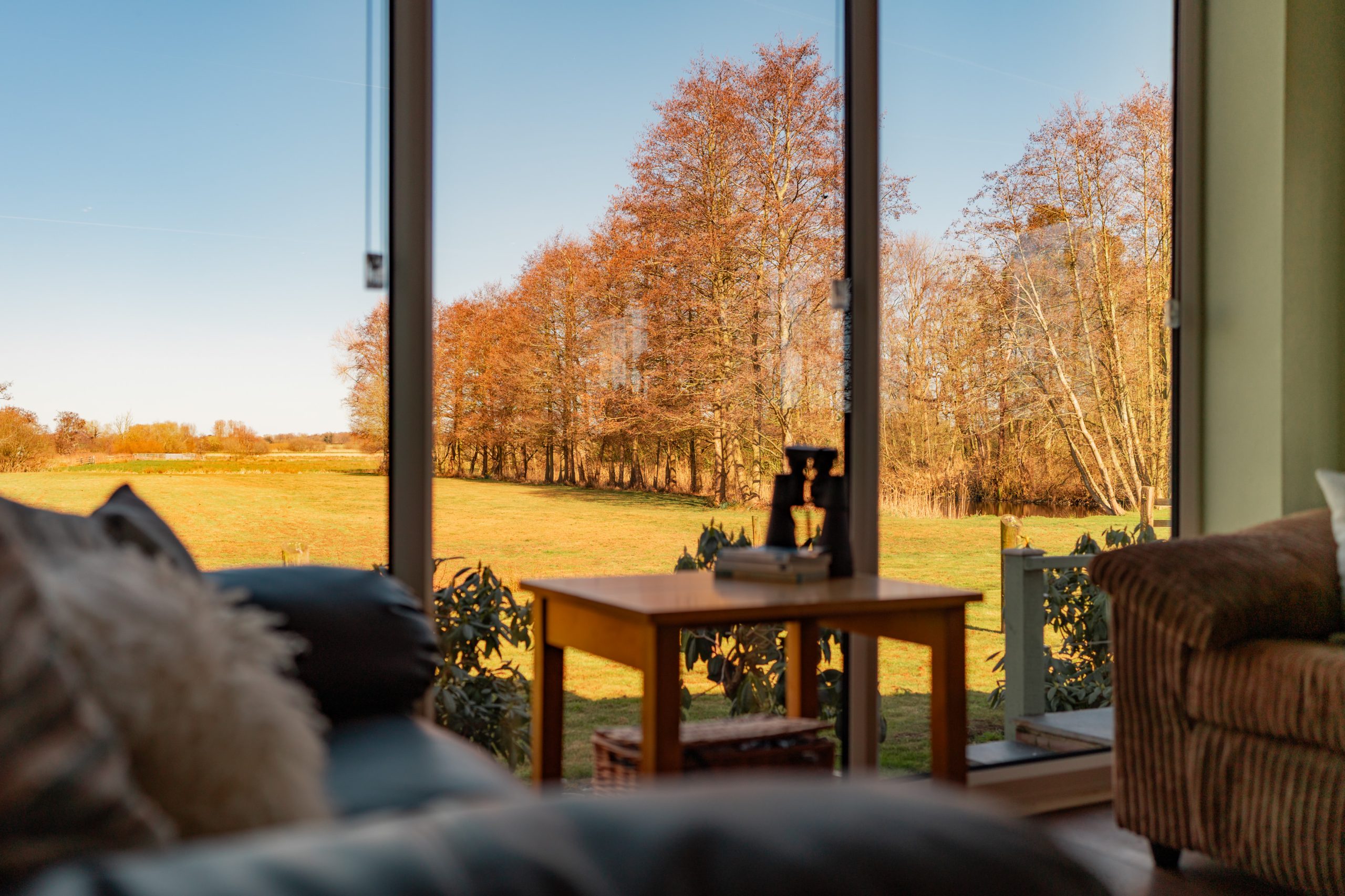A cozy living room with large glass windows overlooks a sunlit autumn landscape. A wooden table with a telescope and books sits by the window. The scene includes a sofa with pillows and views of trees with fall foliage against a clear blue sky.