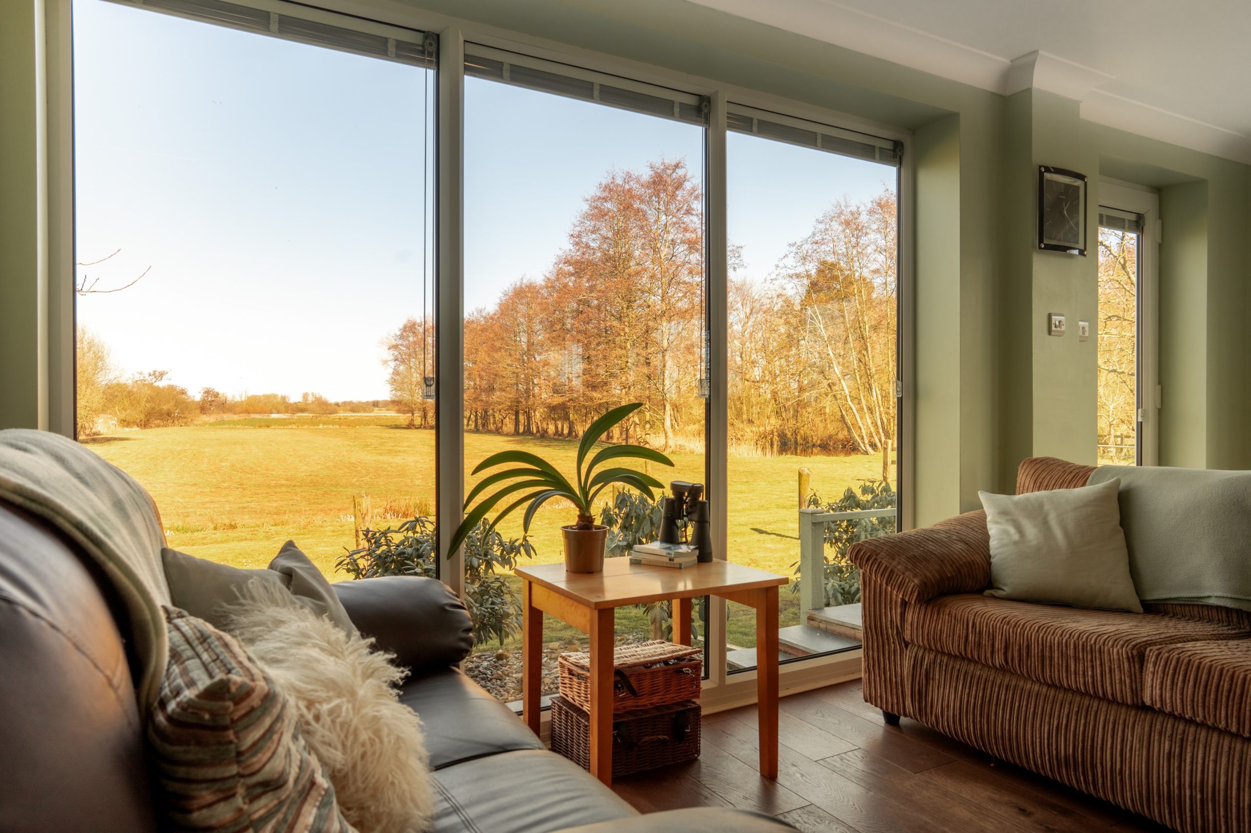 Cozy living room with large windows displaying a scenic view of a field and trees under a clear sky. The room features comfortable sofas, a small wooden table, and a few plants, creating a warm and inviting atmosphere.