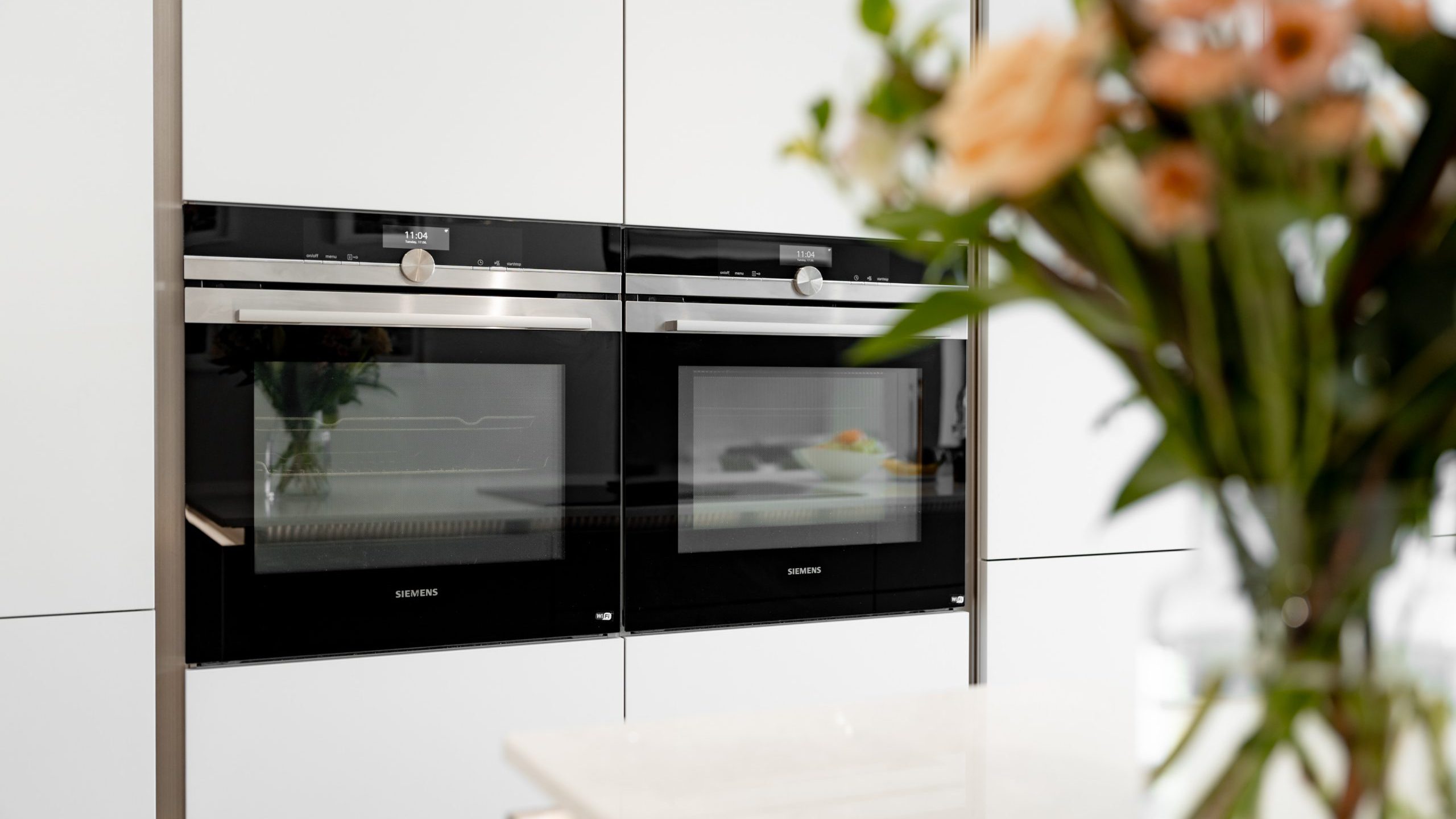 Two built-in Siemens ovens are mounted side by side in a modern white kitchen. A blurred bouquet of flowers is visible in the foreground on the right side.