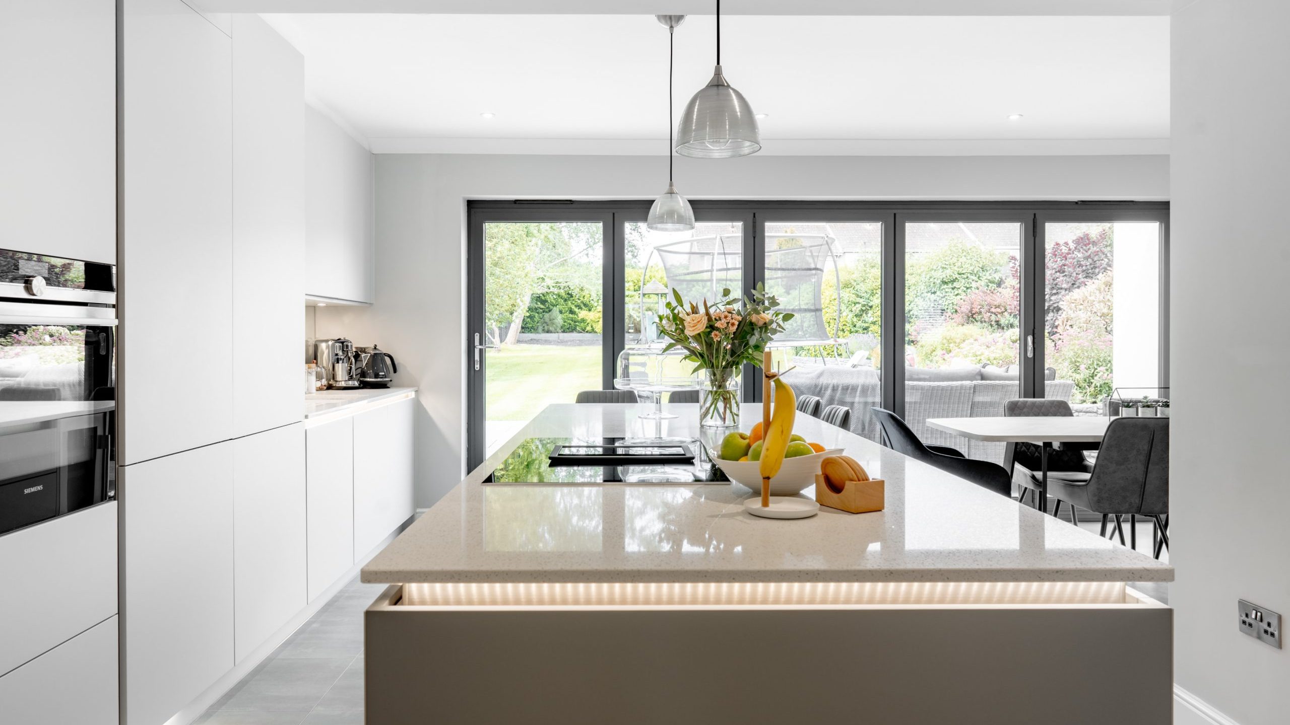 Modern white kitchen with a large island, pendant lights, and fruit bowls. Floor-to-ceiling glass doors open to a green garden and an outdoor patio. Dining area with a table and chairs is visible in the background.