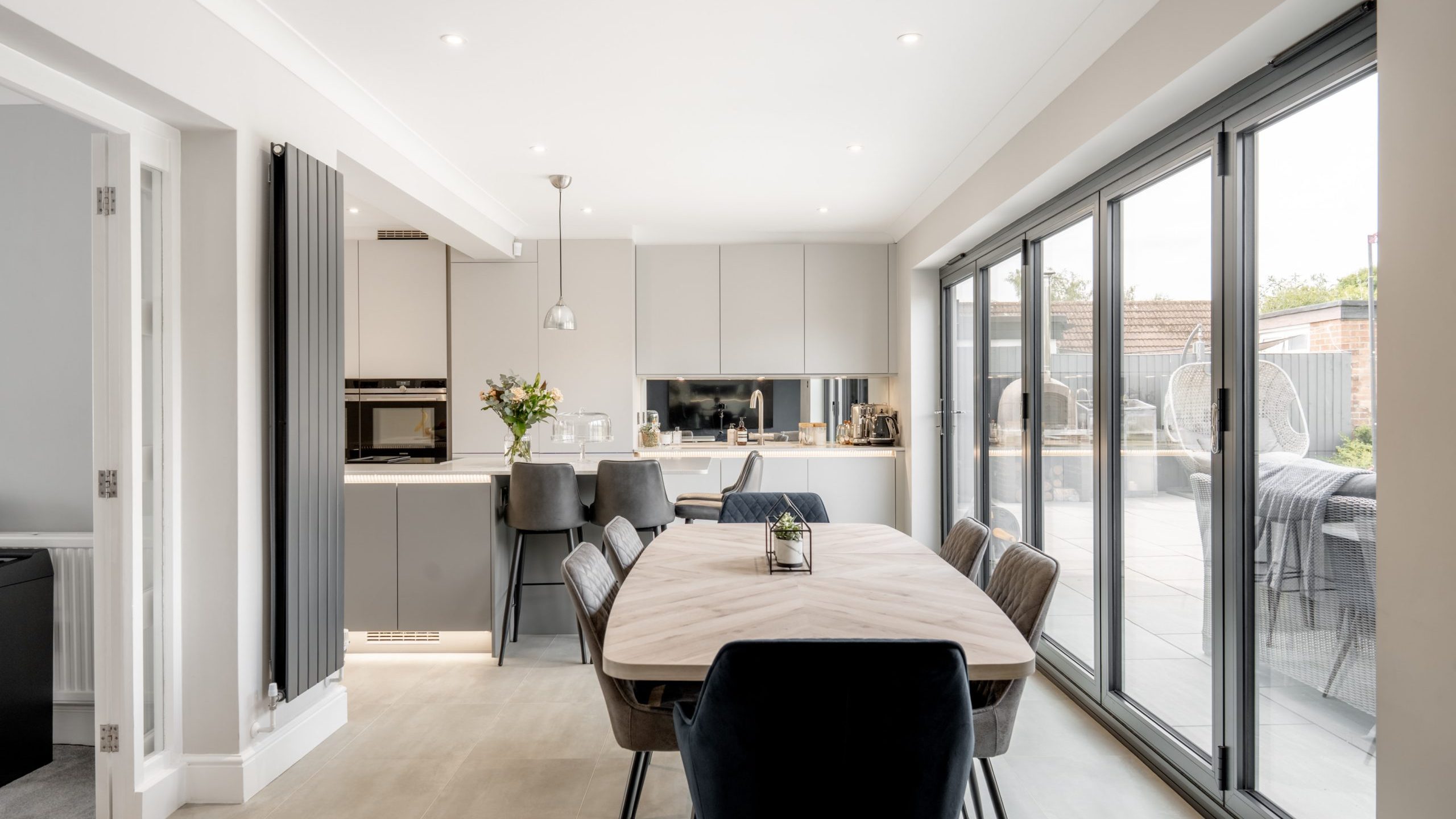 Modern kitchen and dining area with neutral tones, sleek cabinets, a wooden dining table with chairs, large glass sliding doors opening to a patio, and minimalist decor including a vase of flowers on the counter.