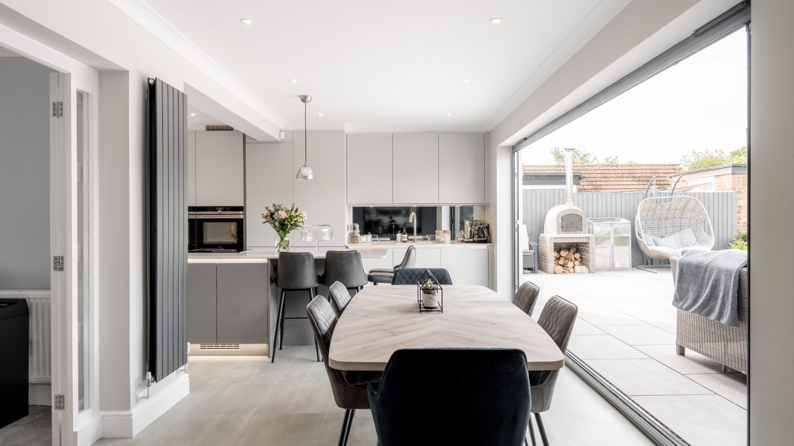 Modern open-plan kitchen and dining area with neutral tones, sleek cabinetry, and a large table with dark chairs. Glass doors open to a bright patio with outdoor seating and a woodpile. Natural light fills the space.