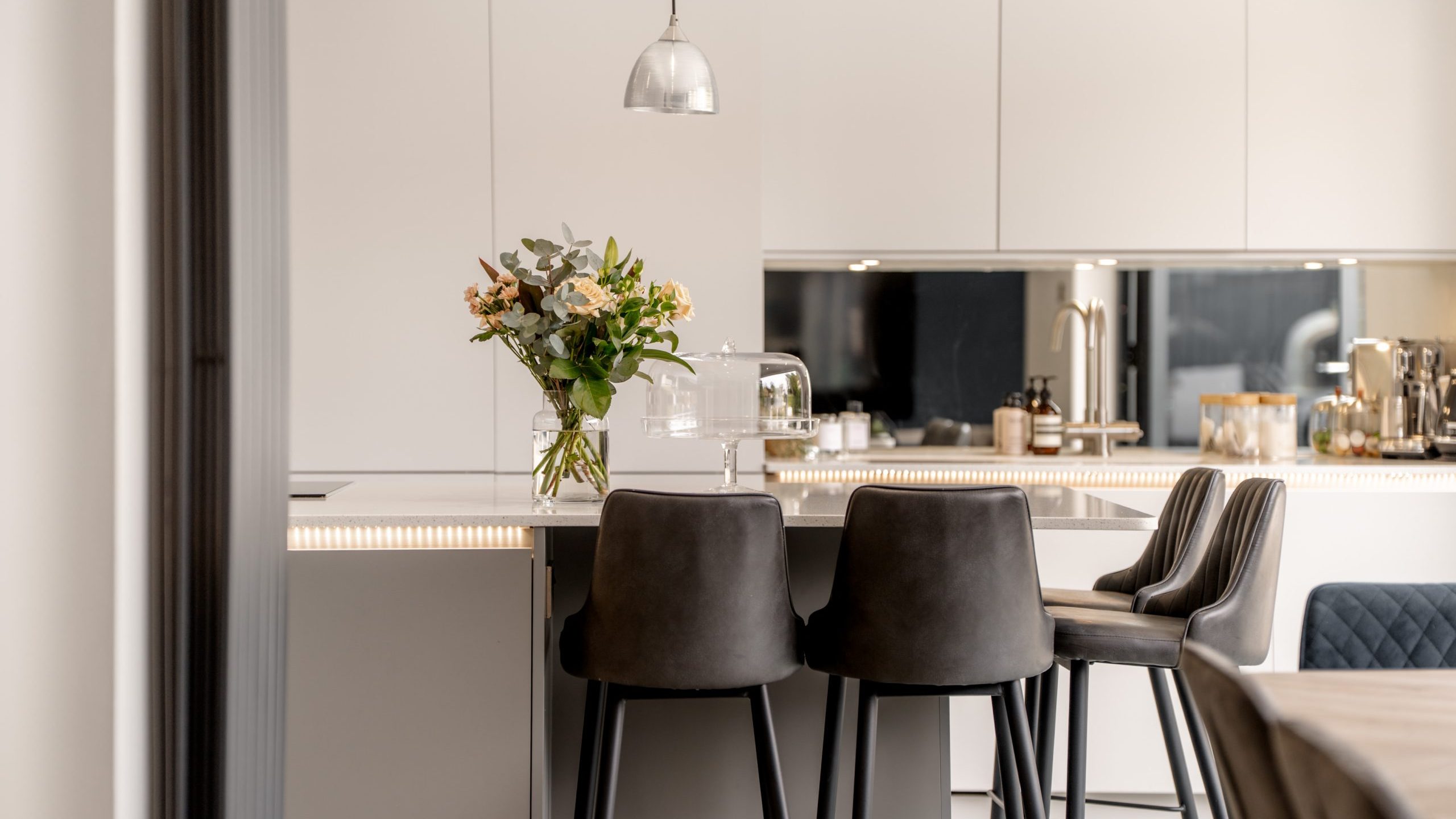 Modern kitchen with white cabinets, a reflective backsplash, and a countertop with a vase of flowers. Three dark gray upholstered bar stools are placed at the counter, beneath a hanging pendant light.