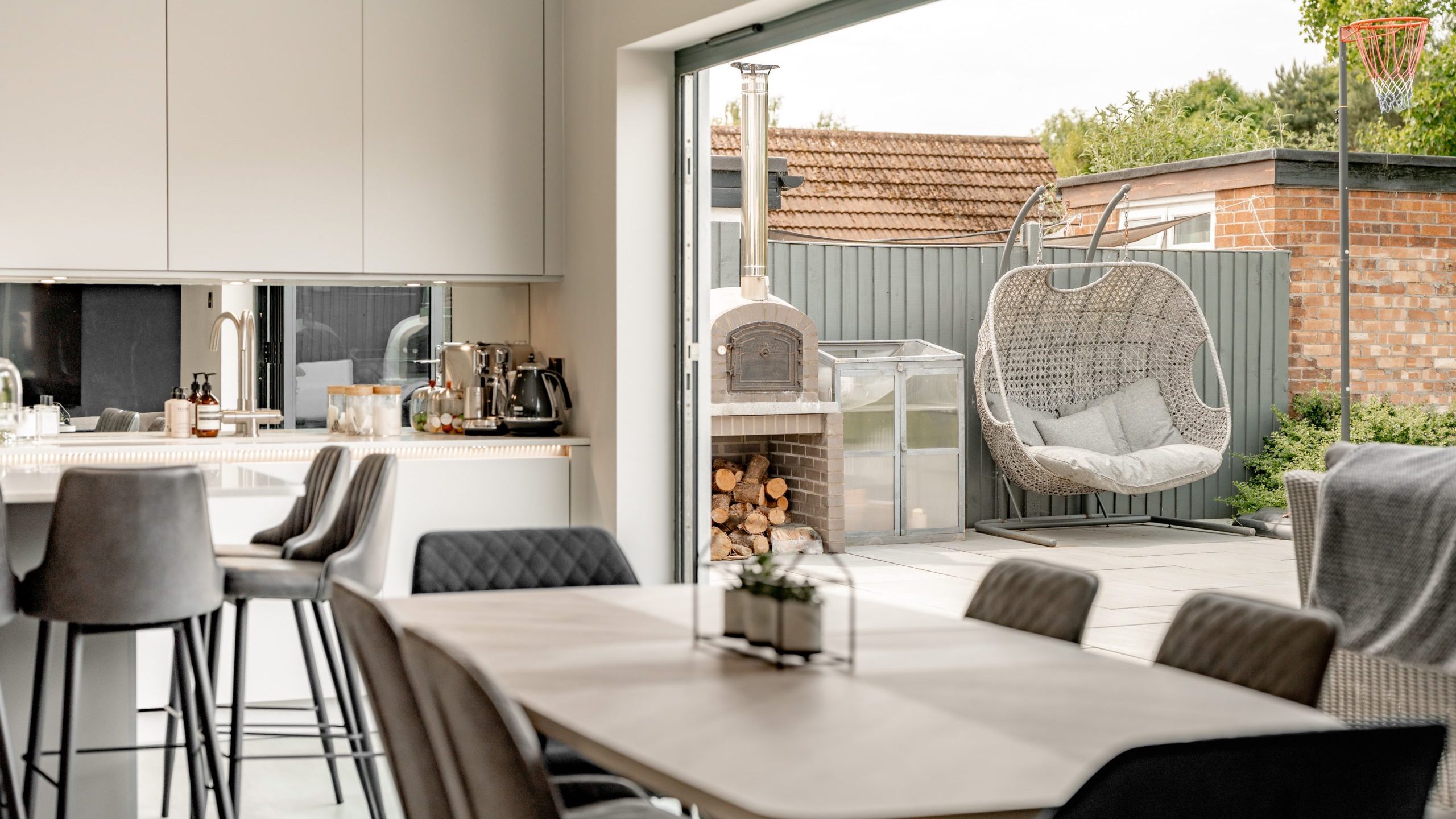 Modern open-plan kitchen and dining area with large sliding doors leading to a patio featuring a hanging egg chair, outdoor pizza oven, stacked firewood, and a basketball hoop in the background.