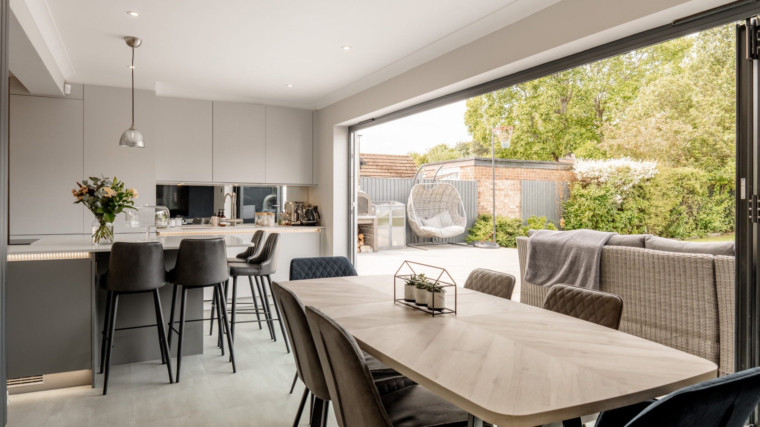 Modern open-plan kitchen and dining area with neutral tones, bar stools at the island, dining table with velvet chairs, and large glass doors opening to a sunny patio with outdoor seating and greenery.