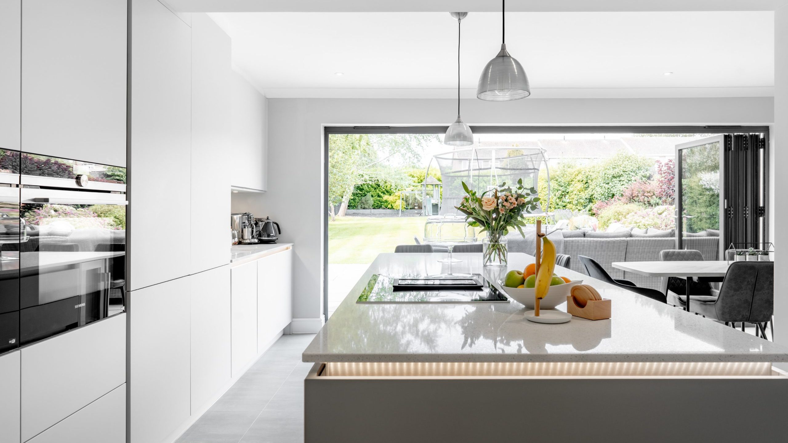 Modern white kitchen with a central island, pendant lights, fresh fruit, and flowers. Large glass doors open to a garden with outdoor seating, bringing in natural light and greenery.