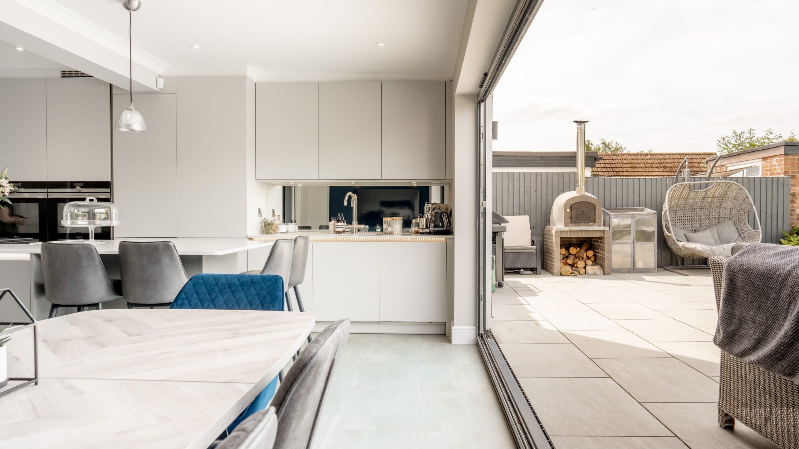 Modern open-plan kitchen and dining area with sliding glass doors leading to a patio with an outdoor oven, log storage, and seating, blending indoor and outdoor living spaces.