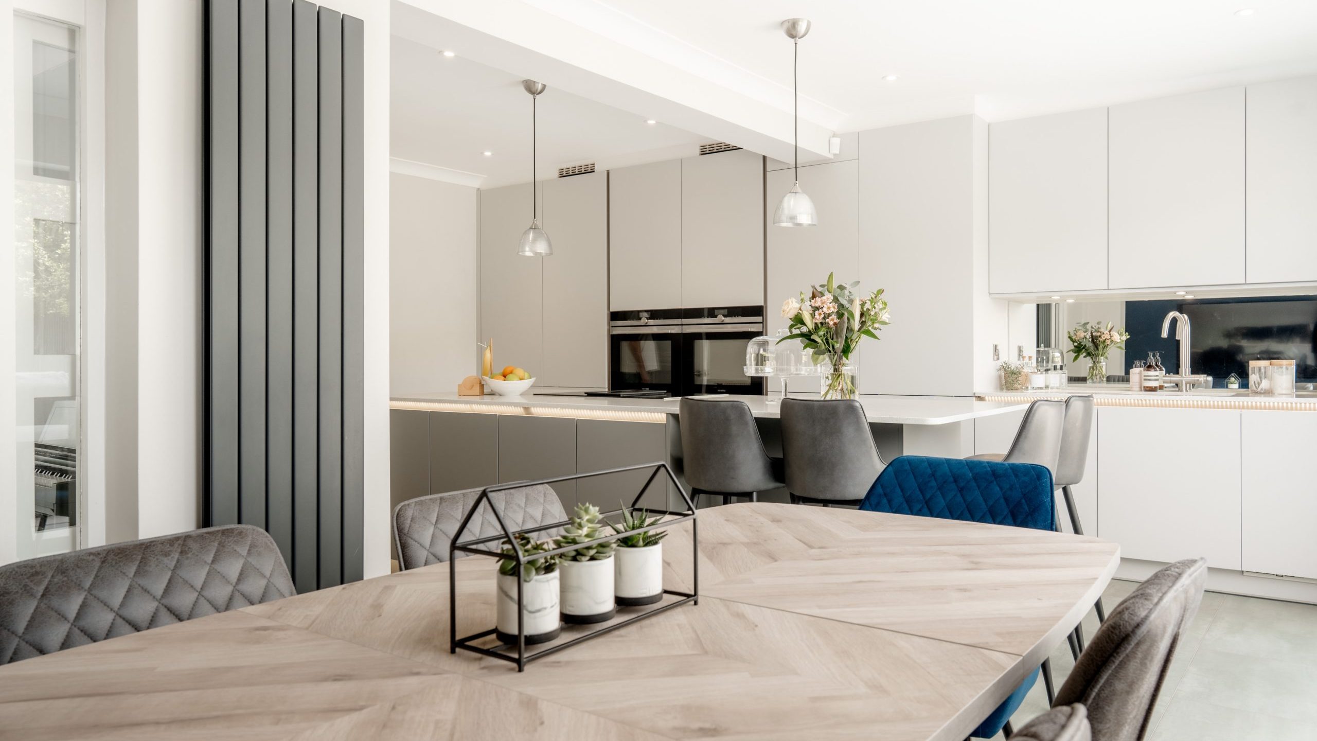 Modern kitchen and dining area with light wood table, gray quilted chairs, potted plants centerpiece, gray cabinets, built-in ovens, pendant lights, and large windows letting in natural light.