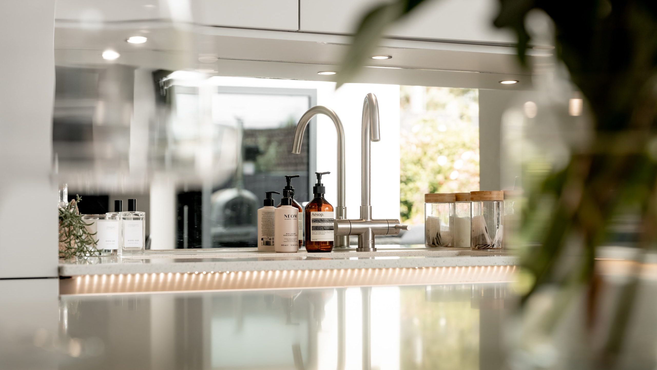 Modern kitchen close-up with a sleek faucet, soap dispensers, and a glass jar on a glossy countertop. Soft lighting and a blurred plant in the foreground create a clean, inviting atmosphere.