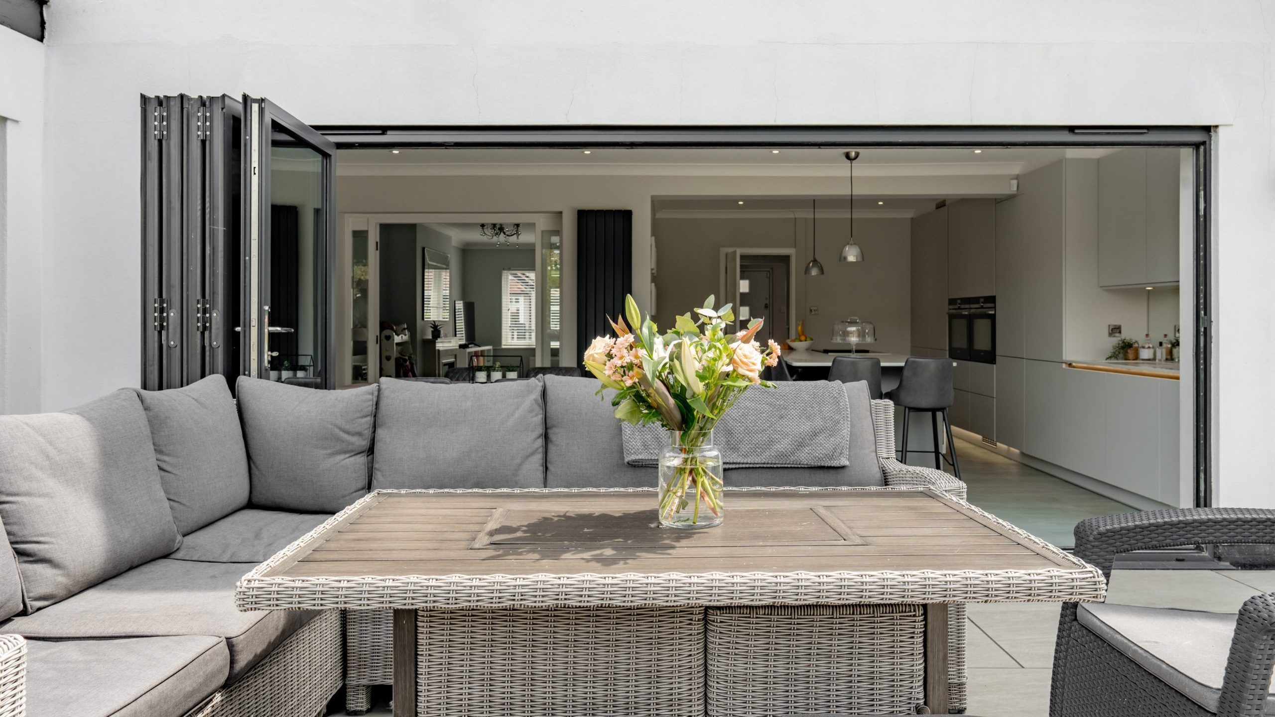 A modern patio with grey wicker furniture and a glass vase of flowers on the table, opening into a spacious kitchen and dining area through large folding glass doors.