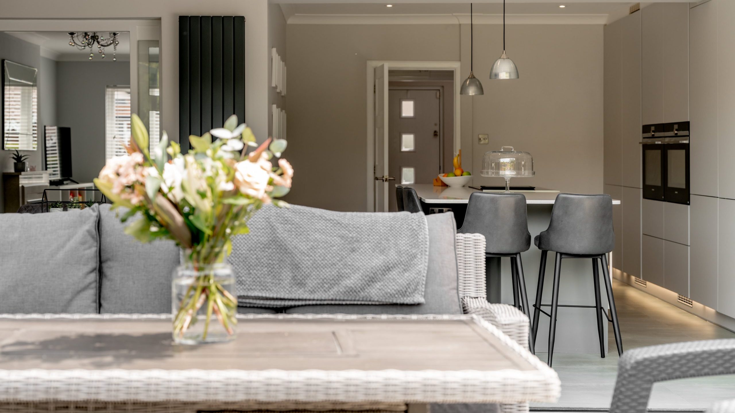 A bright, modern open-plan kitchen and living area with a gray sofa, a vase of flowers on a wicker table, high stools at a kitchen island, and neutral-colored walls and decor.