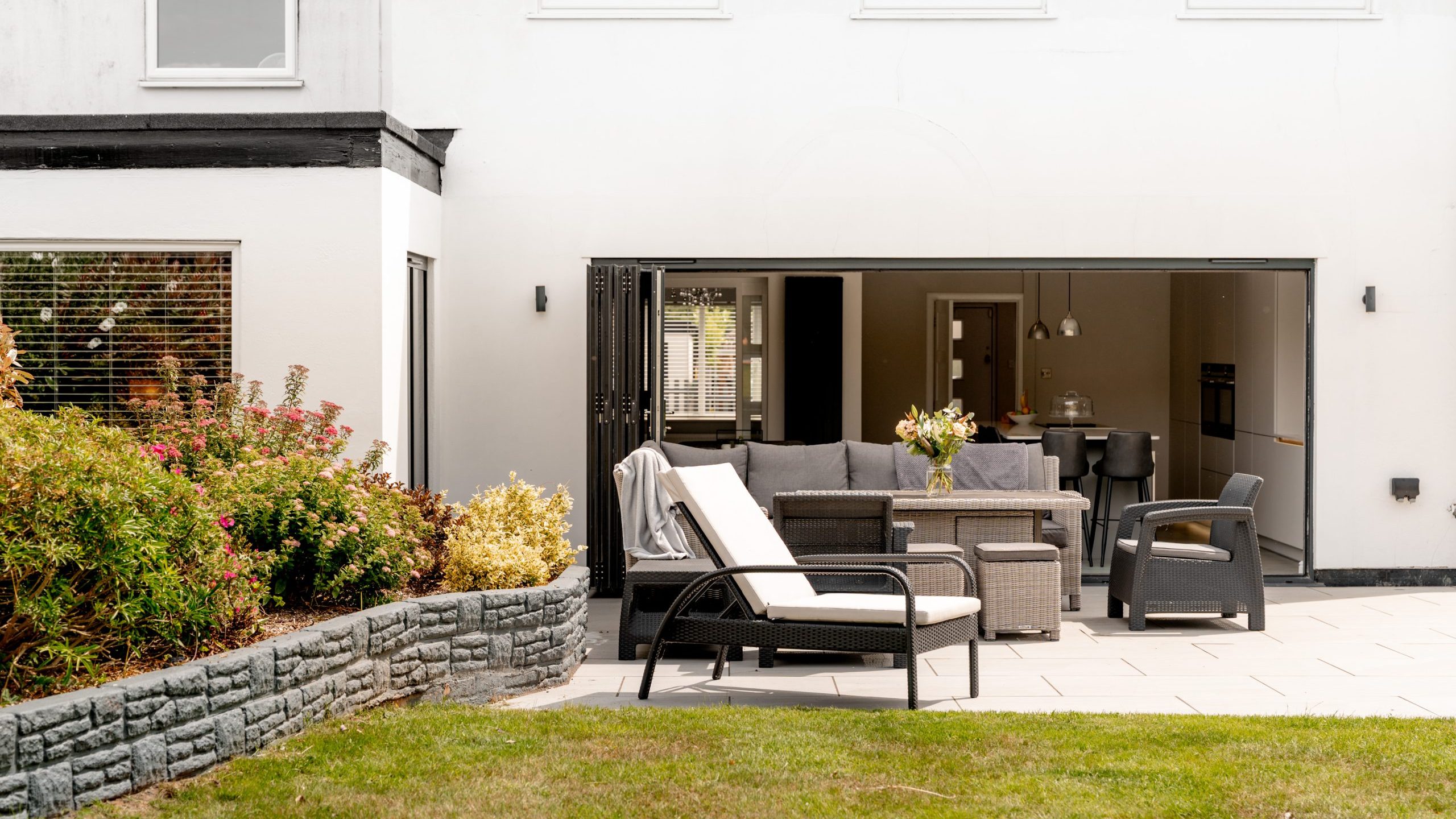 Modern patio with outdoor furniture—lounge chairs, a sofa, and a table—on a paved area by a house with large glass doors opening to a kitchen and dining space. There are green grass and flowering plants nearby.