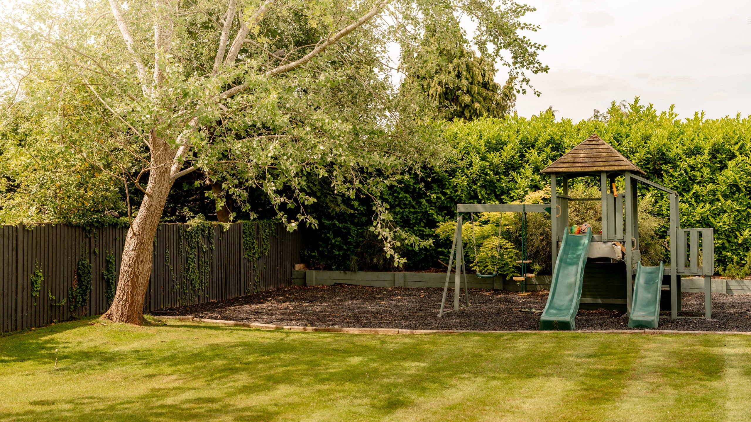 A backyard with green grass, a wooden fence, and a leafy tree. There is a playground set with two green slides, a small climbing structure, and swings, surrounded by wood chips. Tall hedges are in the background.