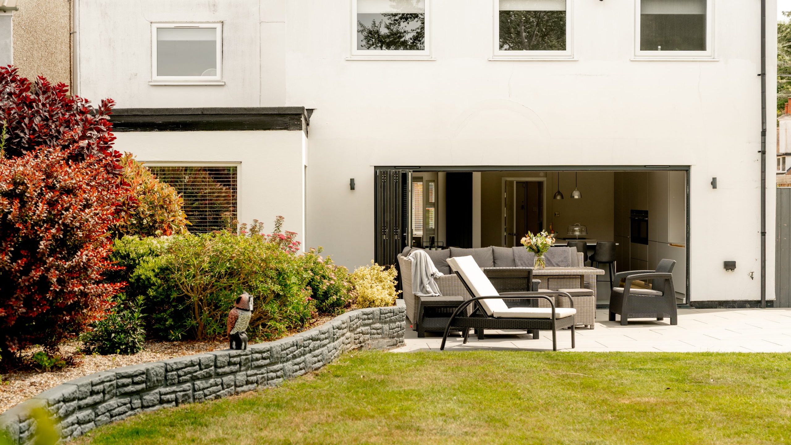 A modern backyard with a patio featuring outdoor furniture, a sofa, and chairs. The lawn is neatly trimmed, and there are shrubs and small trees along a stone-edged garden bed next to a white house with large windows.