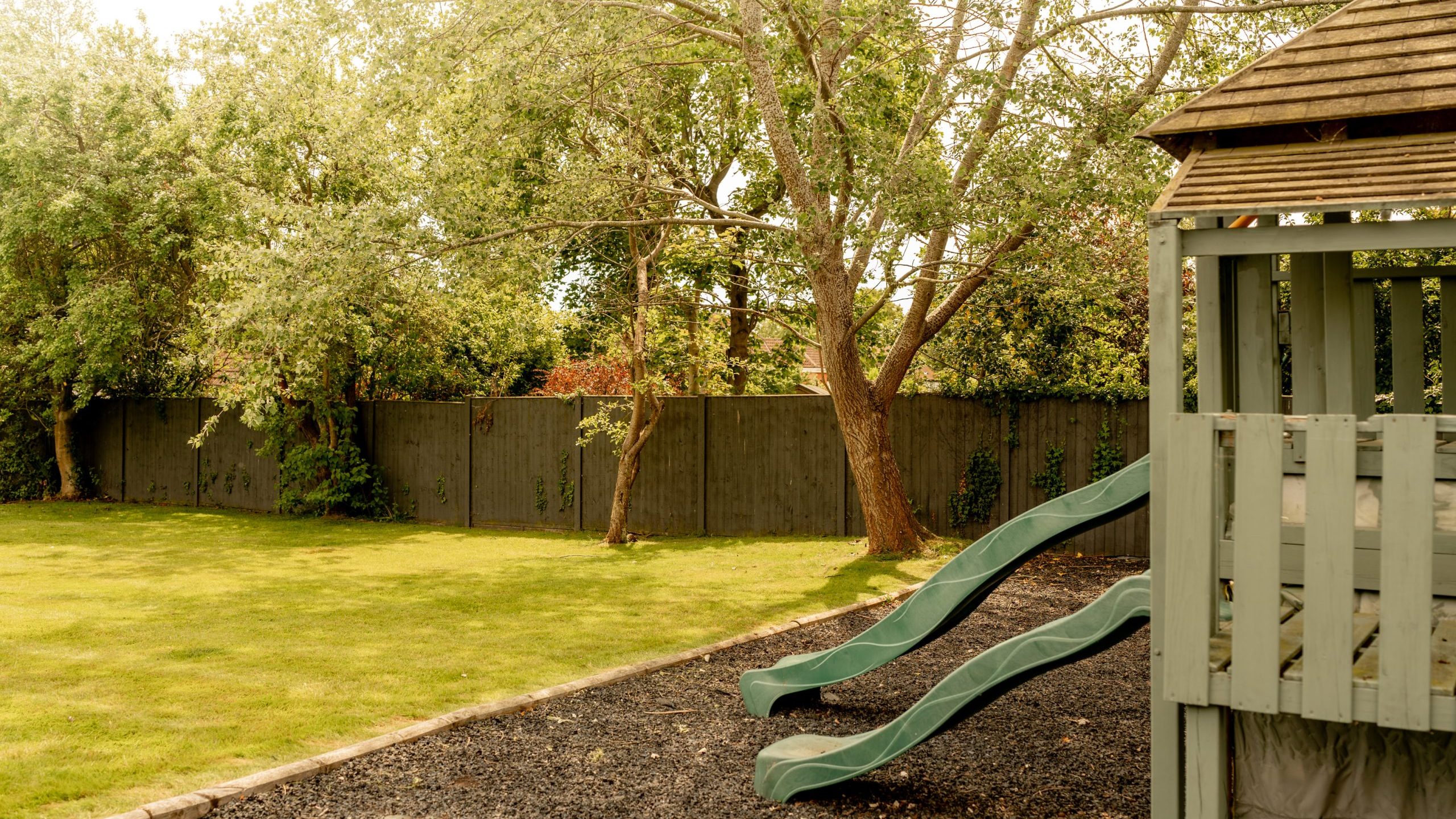 A backyard with green grass, tall trees, a wooden fence, and a play structure featuring two green slides on the right side. The scene is bright and sunny.