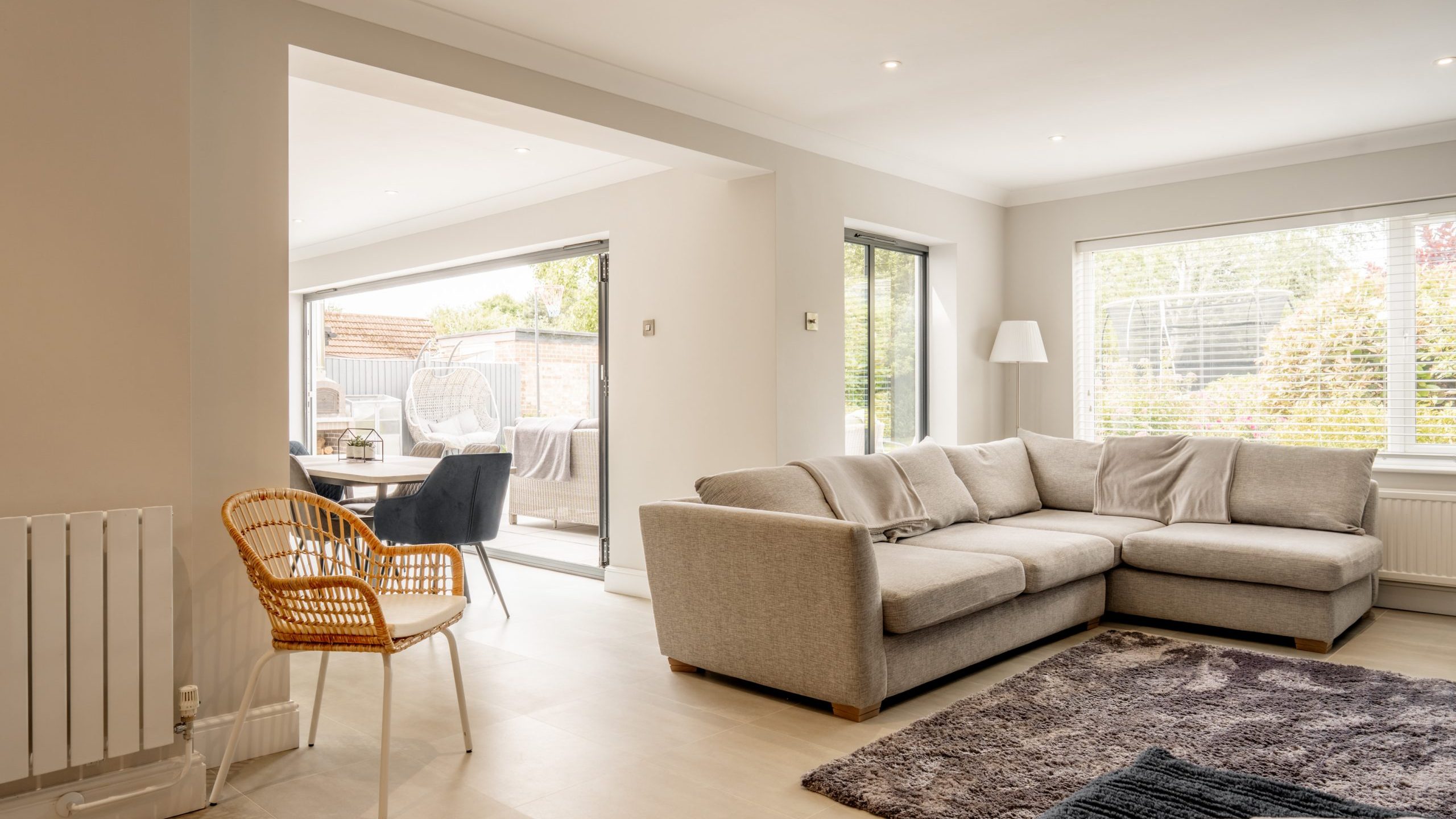 Bright, modern living room with a large L-shaped gray sofa, a wicker chair, a gray rug, and large windows allowing natural light in. The adjoining dining area is visible with sliding glass doors to a patio.