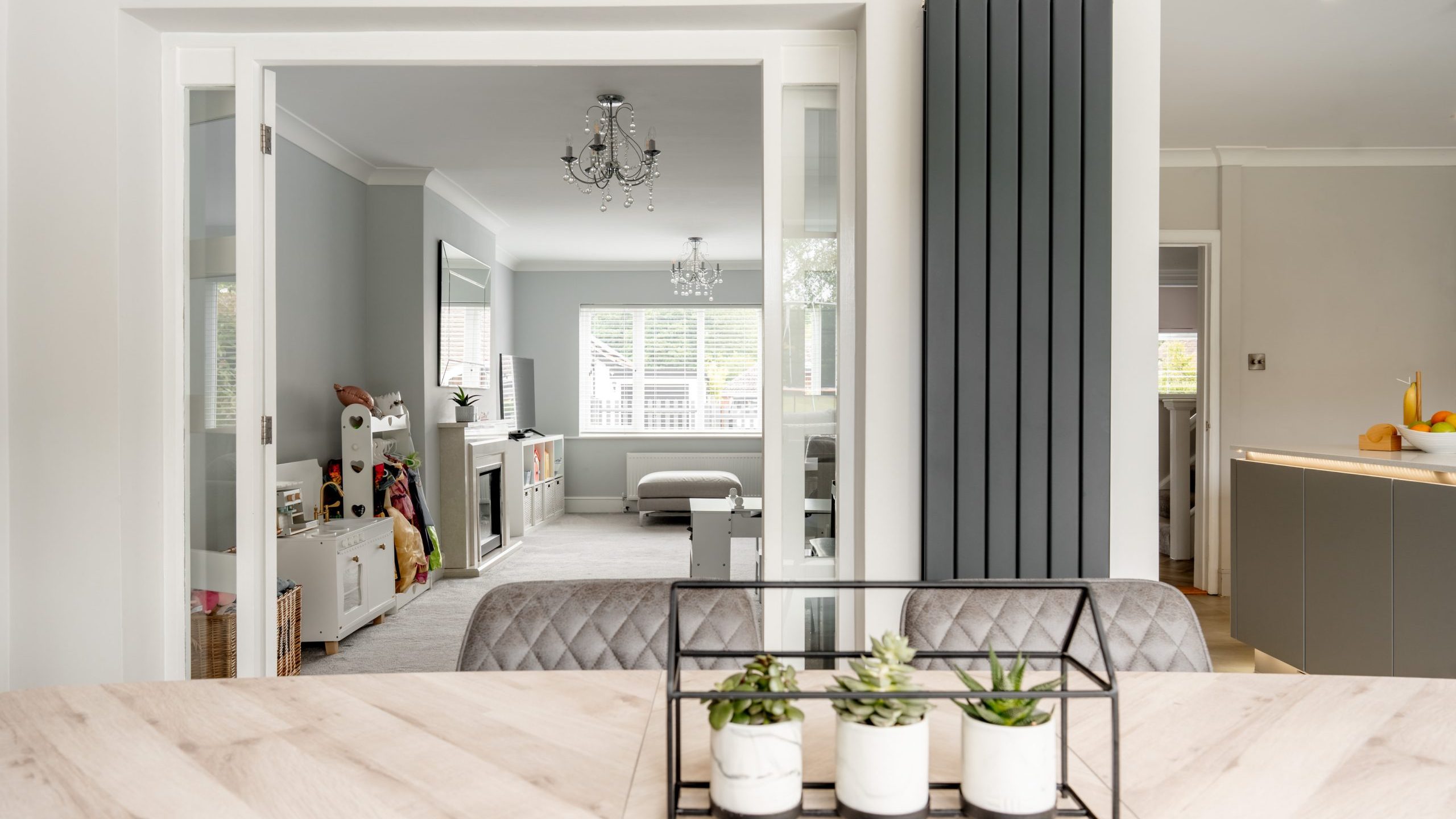 A modern, bright living space with large windows, neutral colors, a wooden dining table with potted succulents, and a view into a cozy room with toys, storage cubes, and a chandelier.