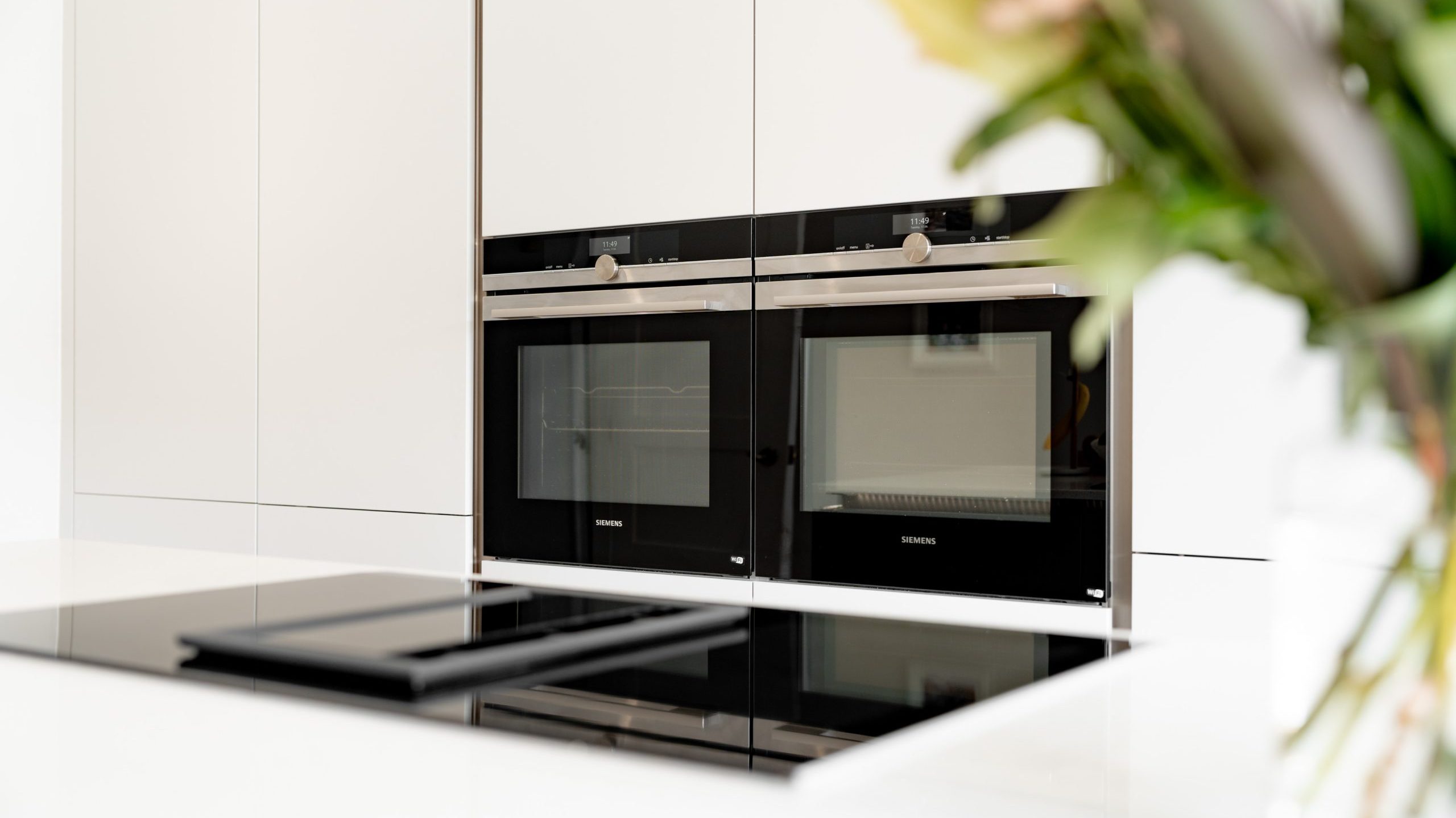 A modern kitchen with built-in Siemens ovens and a black glass induction cooktop, featuring white cabinetry and blurred green and pink flowers in the foreground.