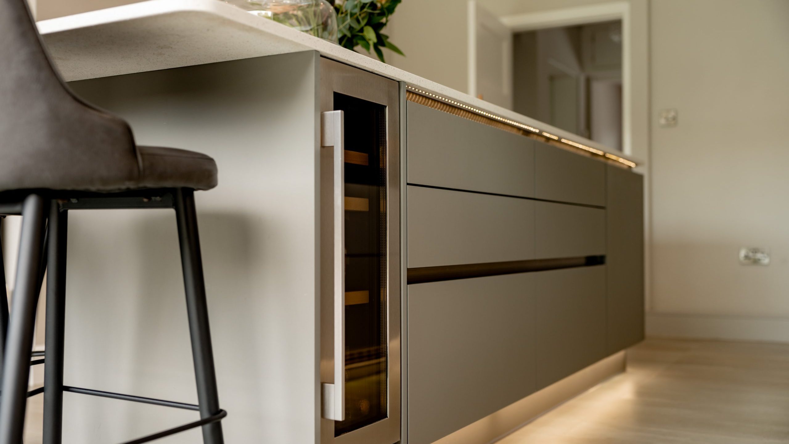 Modern kitchen island with matte gray drawers, built-in wine cooler, and a light countertop. A dark bar stool is beside the island, and a vase with flowers sits on top. The background shows a doorway and soft lighting.