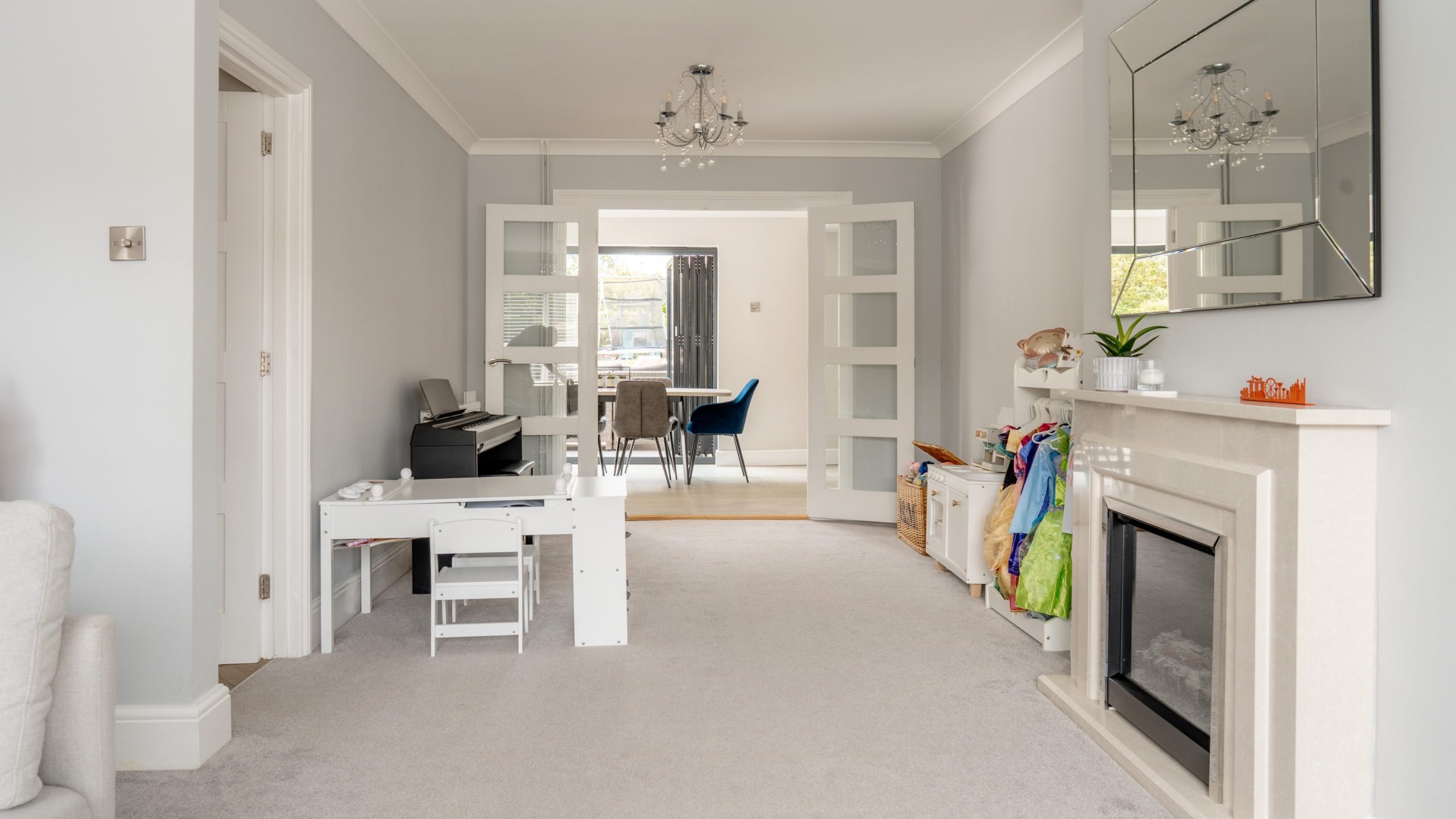 A bright, modern living room with light gray walls, a fireplace, a large mirror, a children’s play area, and an upright piano. Double doors open to a dining room with a table and blue chairs in the background.
